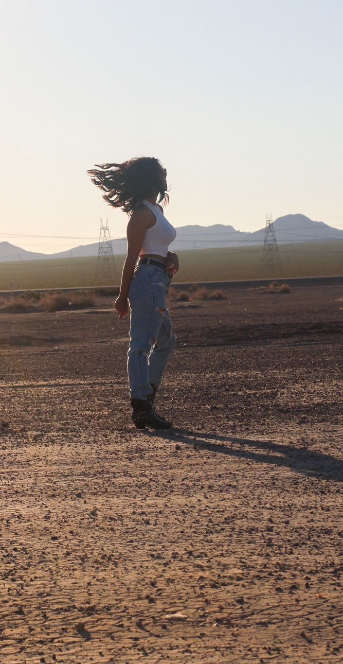 Woman in white crop top and jeans with wind-blown hair standing in vast desert plain.