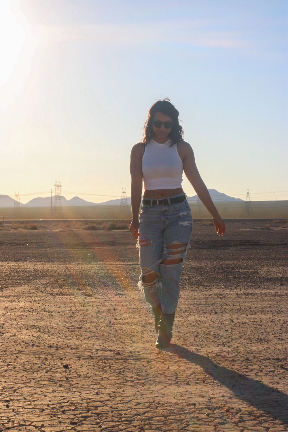 Woman in white crop top and ripped jeans walking in sunlit cracked desert landscape.