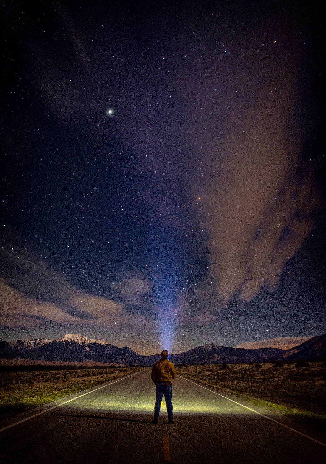 Silhouette of person standing on road under starry night sky with mountains in background.