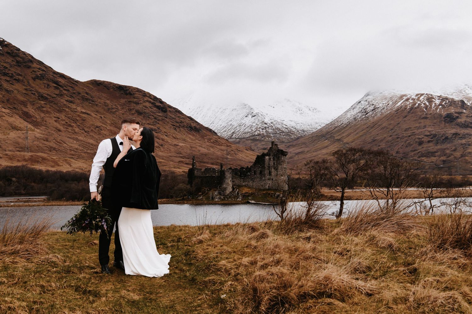 Couple embracing in a scenic Scottish Highland landscape with mountains and a loch in the background.