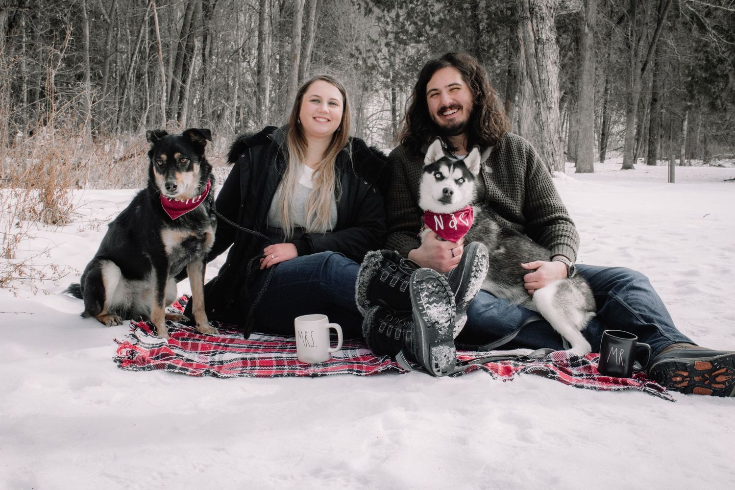Couple sitting on plaid blanket in snow with two husky dogs.