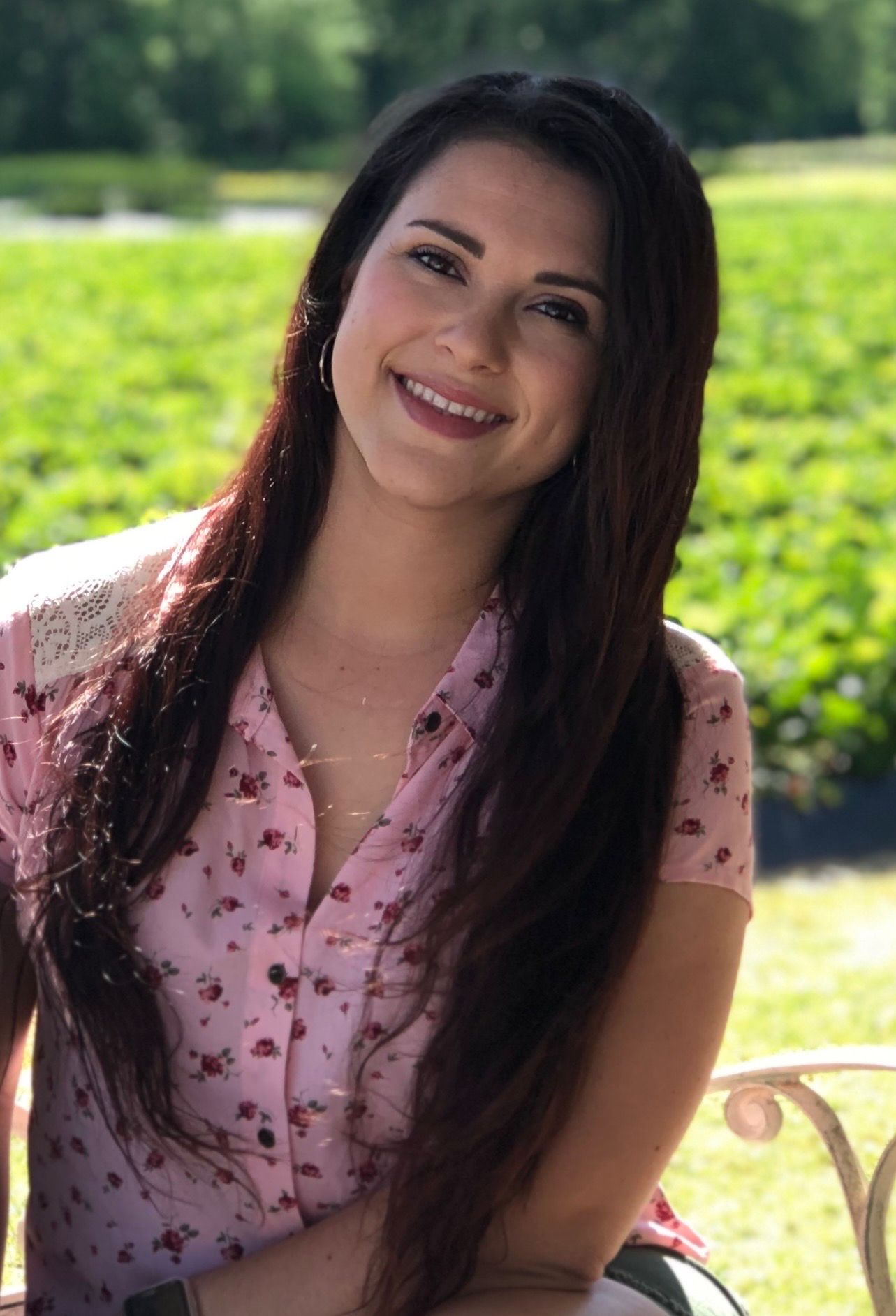 Model in pink floral dress poses outdoors in sunny garden setting.