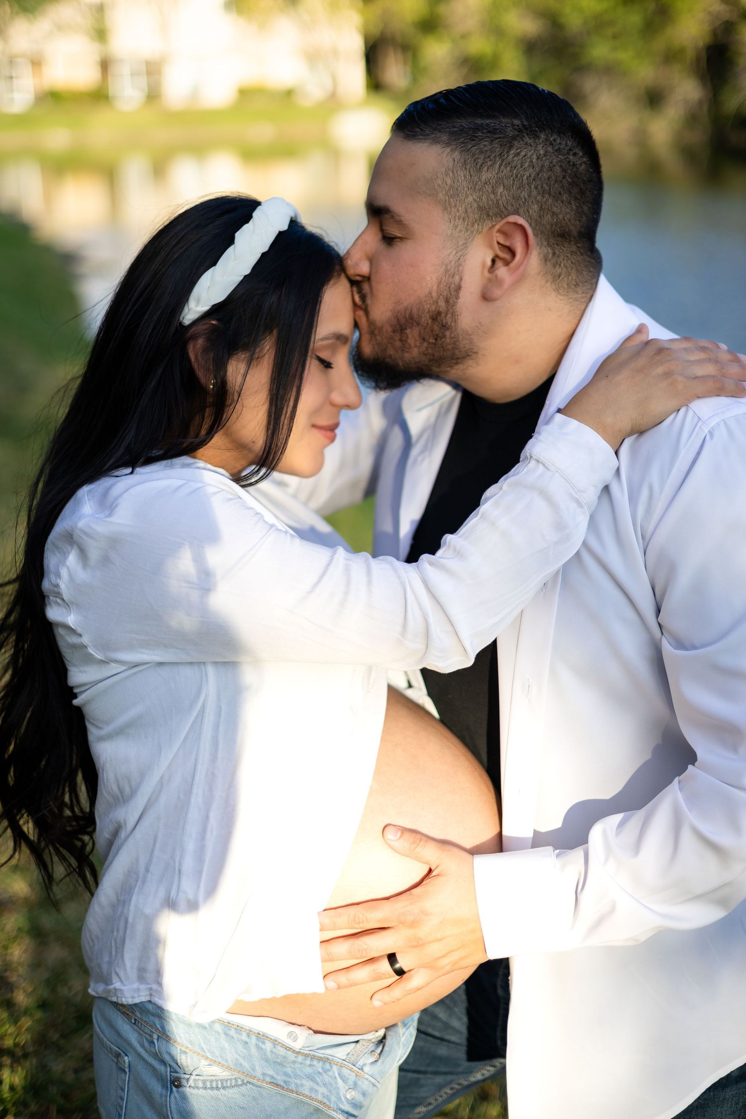 An expectant couple sharing an intimate moment outdoors near water.