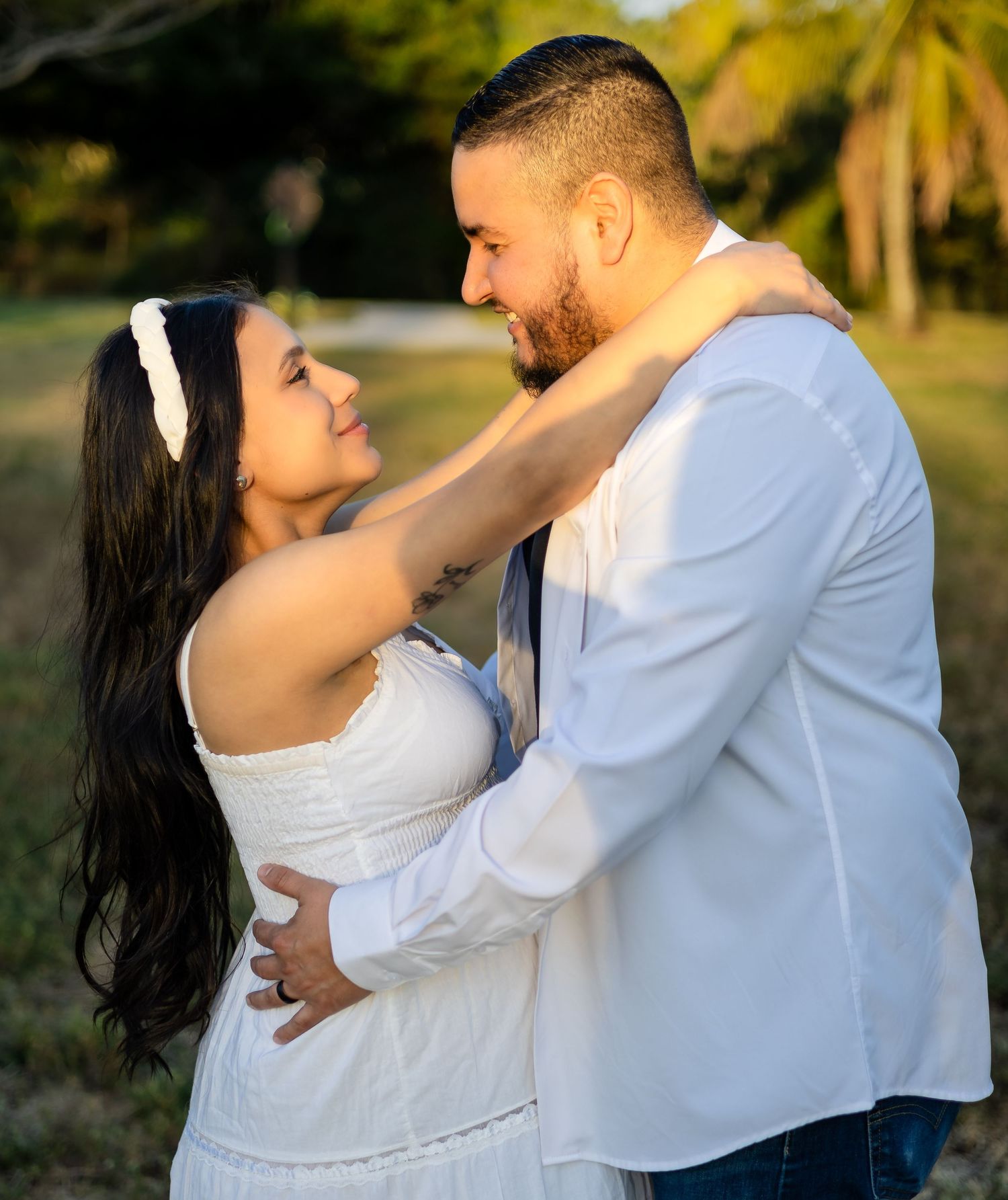 A romantic couple embraces outdoors in white clothing with palm trees in the background during golden hour.