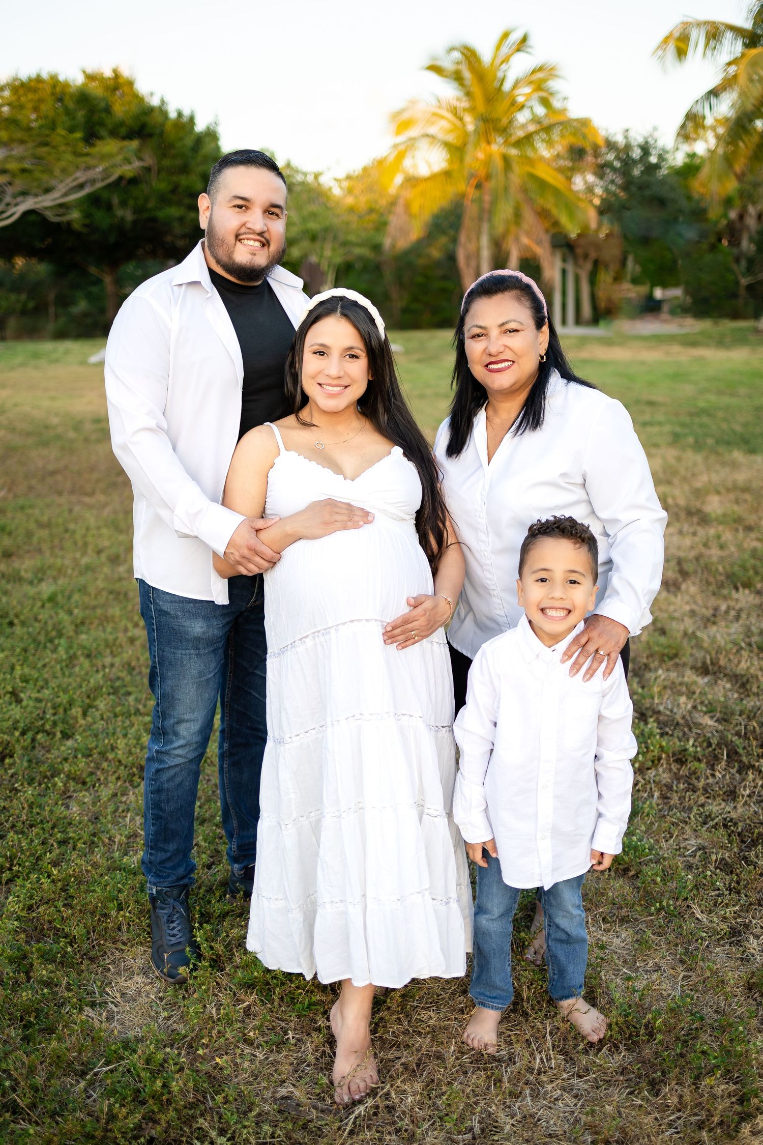 A family of four poses together outdoors for a maternity portrait with palm trees behind them.