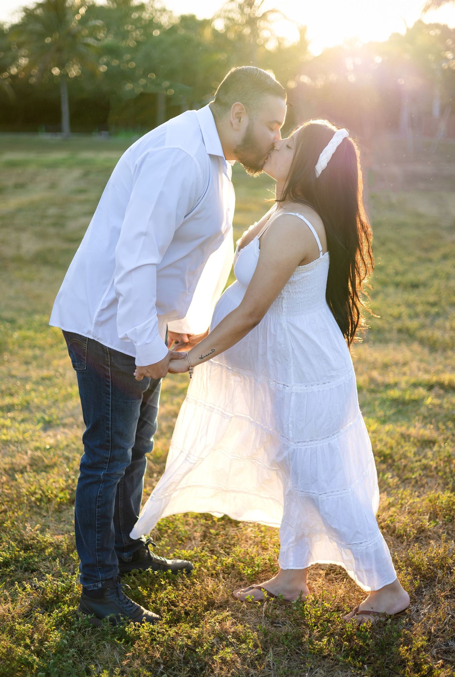 A romantic sunset maternity photo of a couple in white clothing against a golden sky.