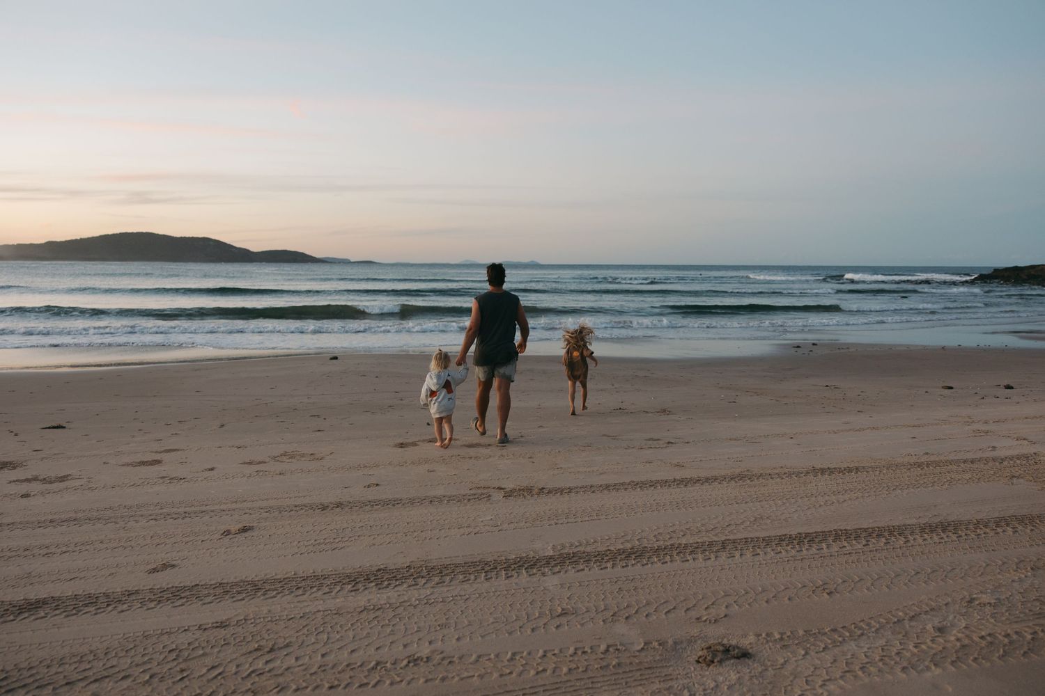 Figure walking dogs along empty beach at sunset with ocean waves in background.