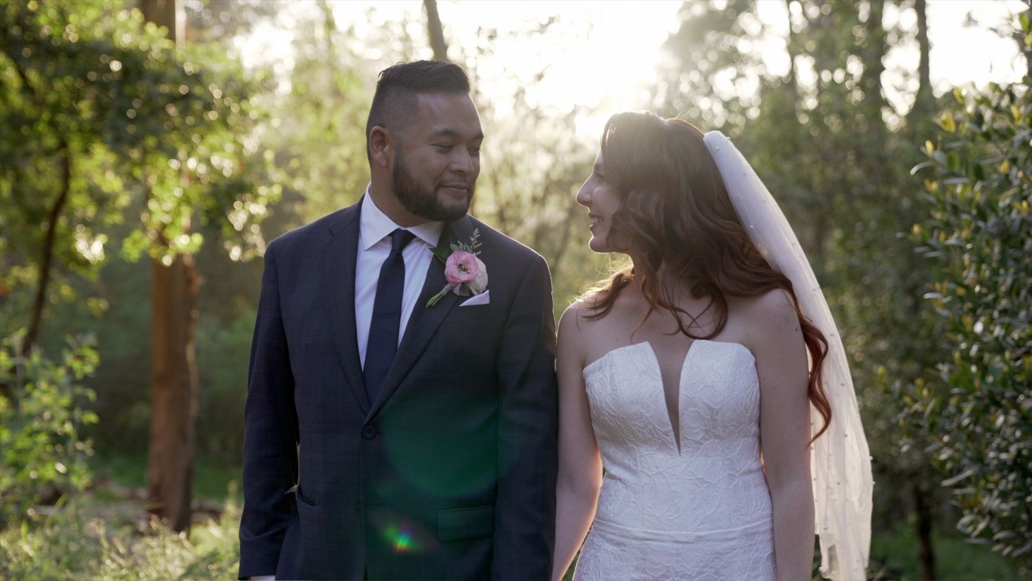 A wedding couple gazes at each other while standing in a sunlit garden with lens flare effects.