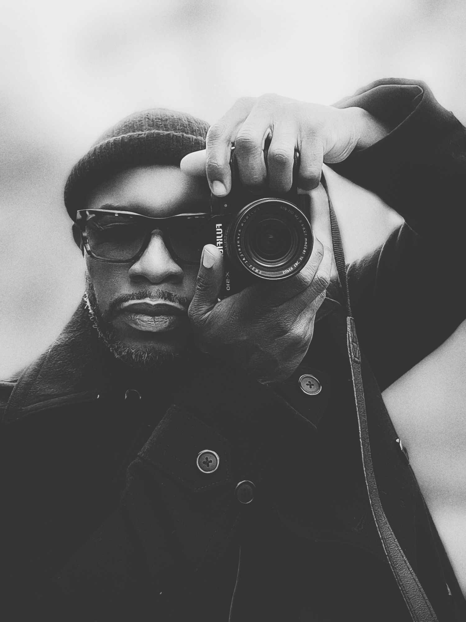 Brian Autry, Black and white portrait of a photographer in sunglasses and coat holding up a camera to taking street photography.