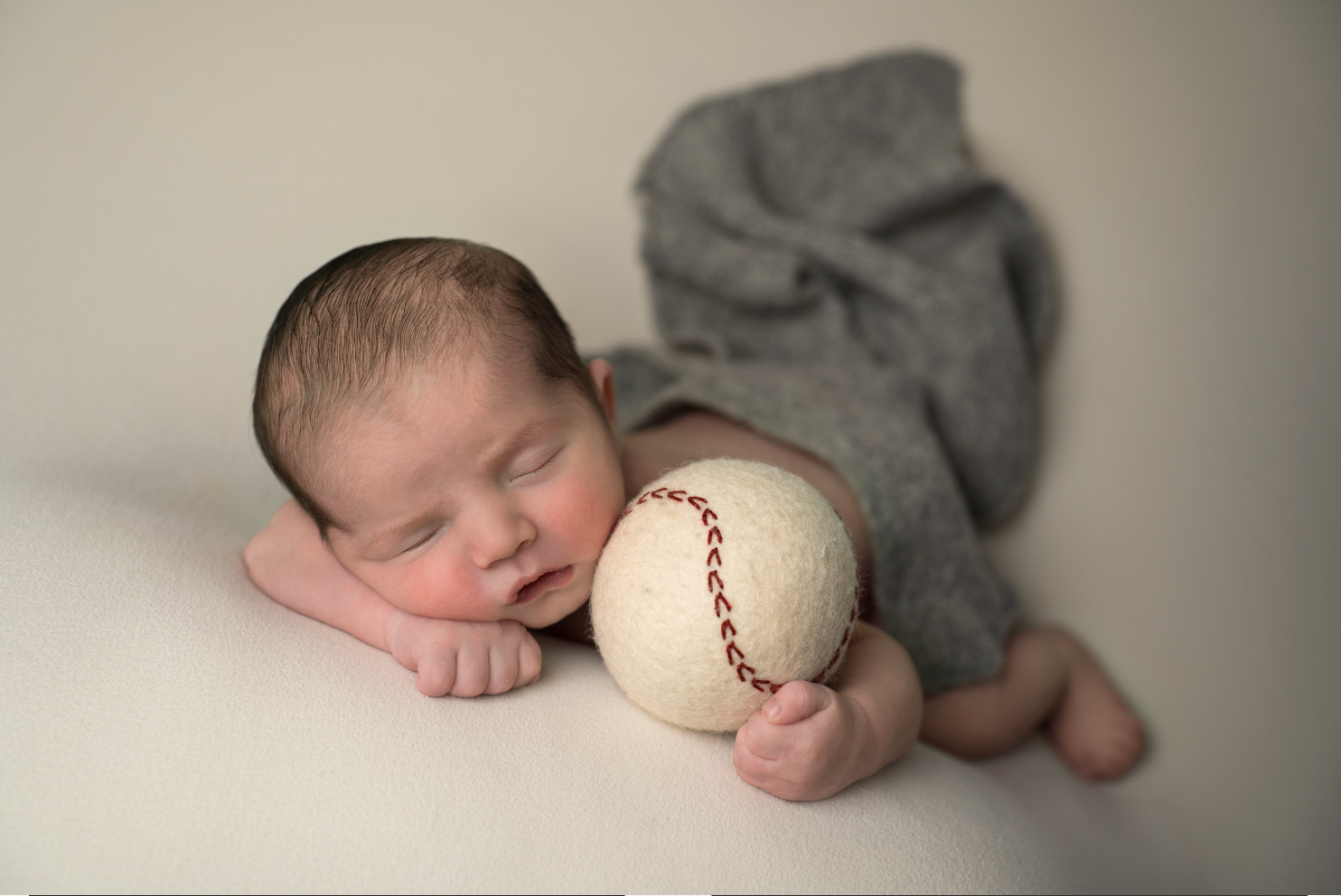 Sleeping newborn baby in gray wrap cuddling a baseball prop during a professional newborn photography session. Embracing Life’s Beauty