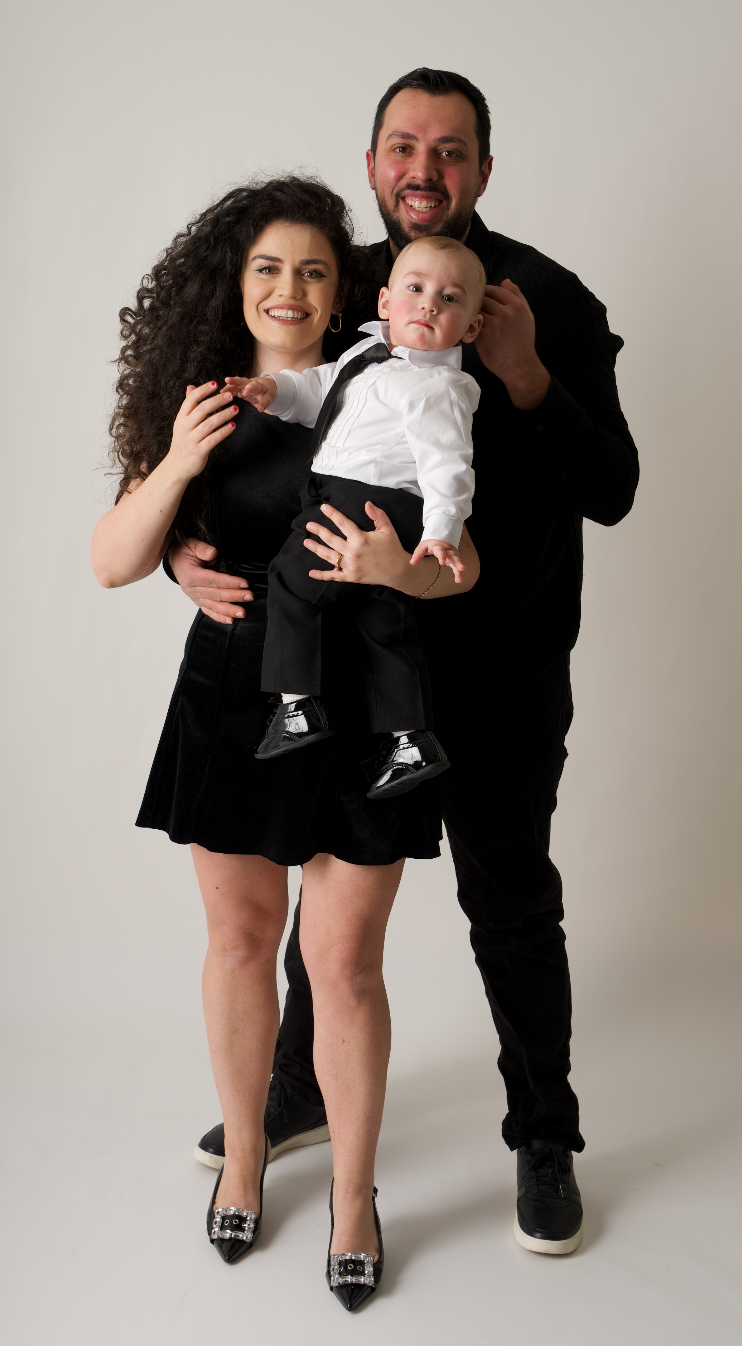 Family of three in matching black outfits, parents holding baby boy in white shirt and tie, studio portrait.
