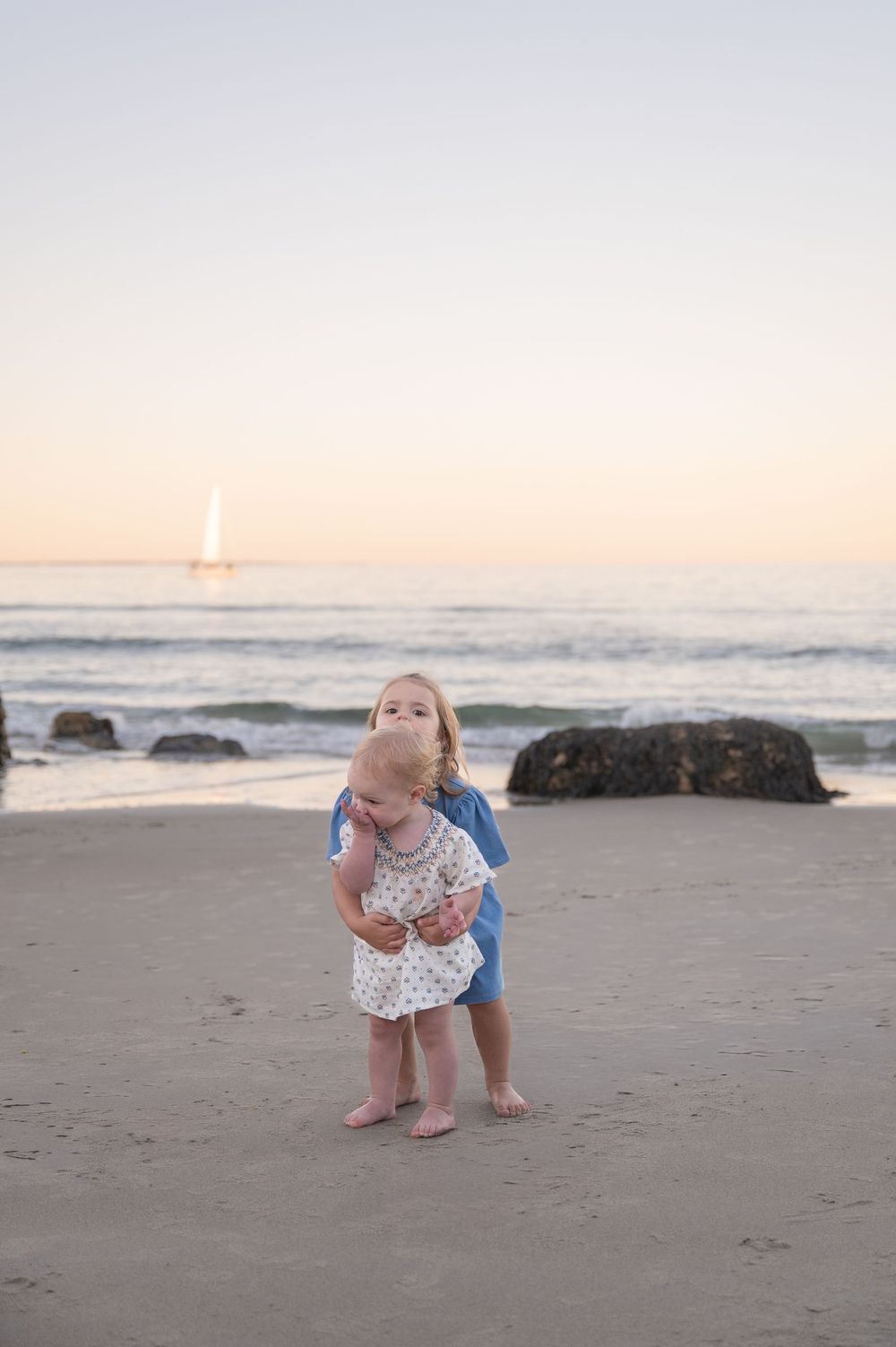 Two young children stand on a sandy beach at sunset with a sailboat visible on the calm ocean horizon.
