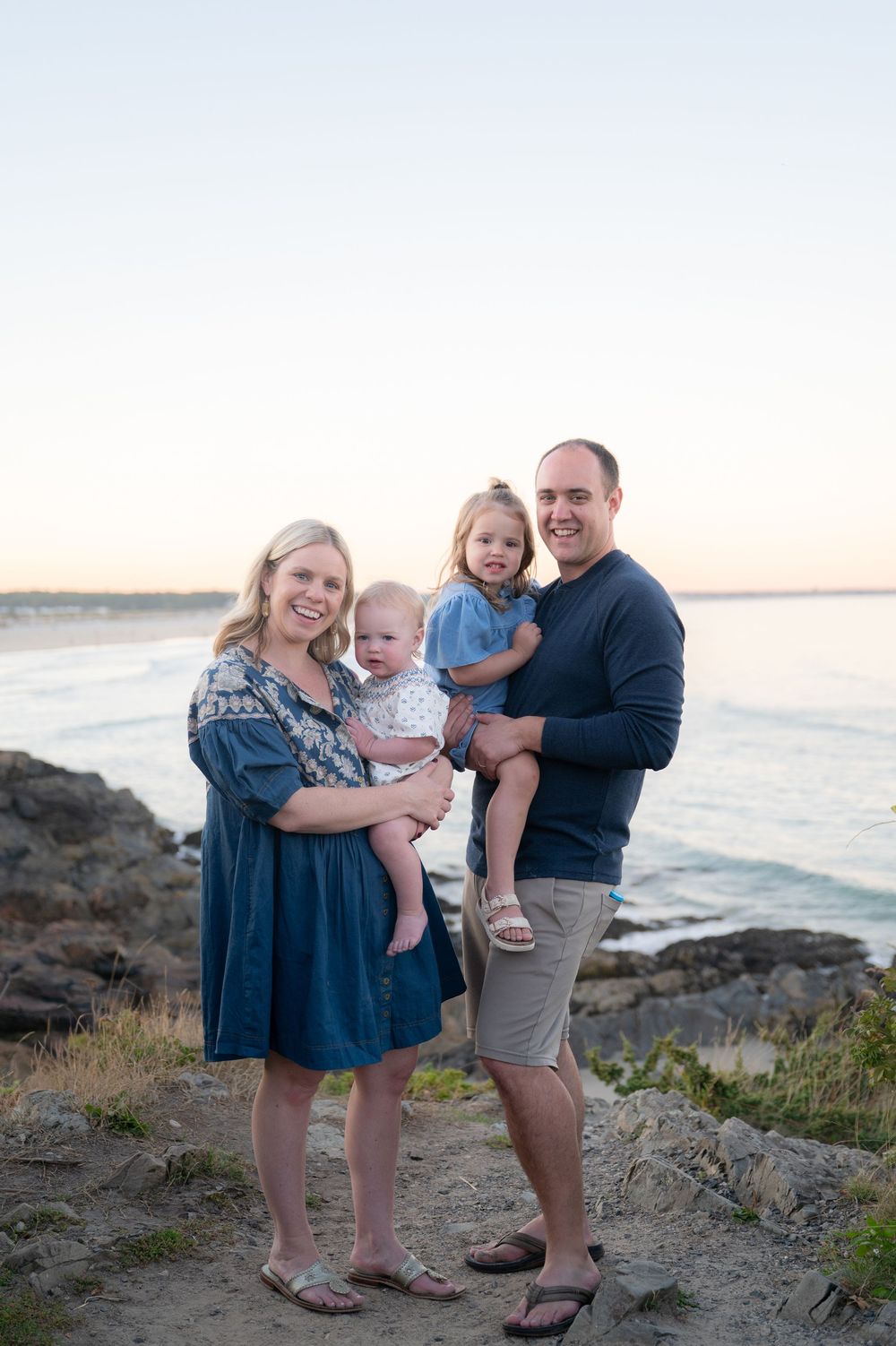 Family of four posing on rocky coastal shore at sunset, parents holding two young children with ocean waves behind them.