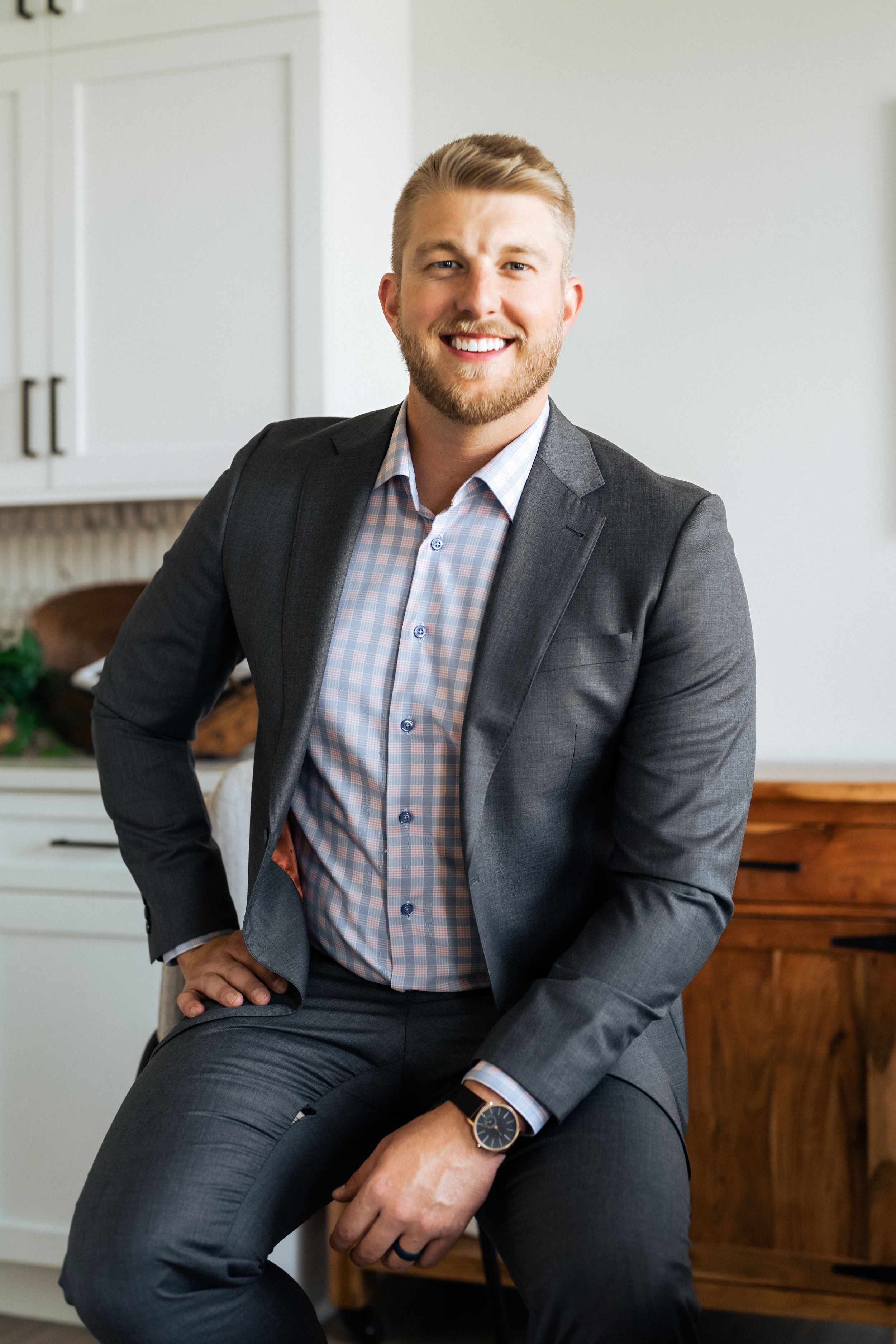 Smiling man in gray blazer and plaid shirt seated in a bright modern kitchen, wearing a watch.