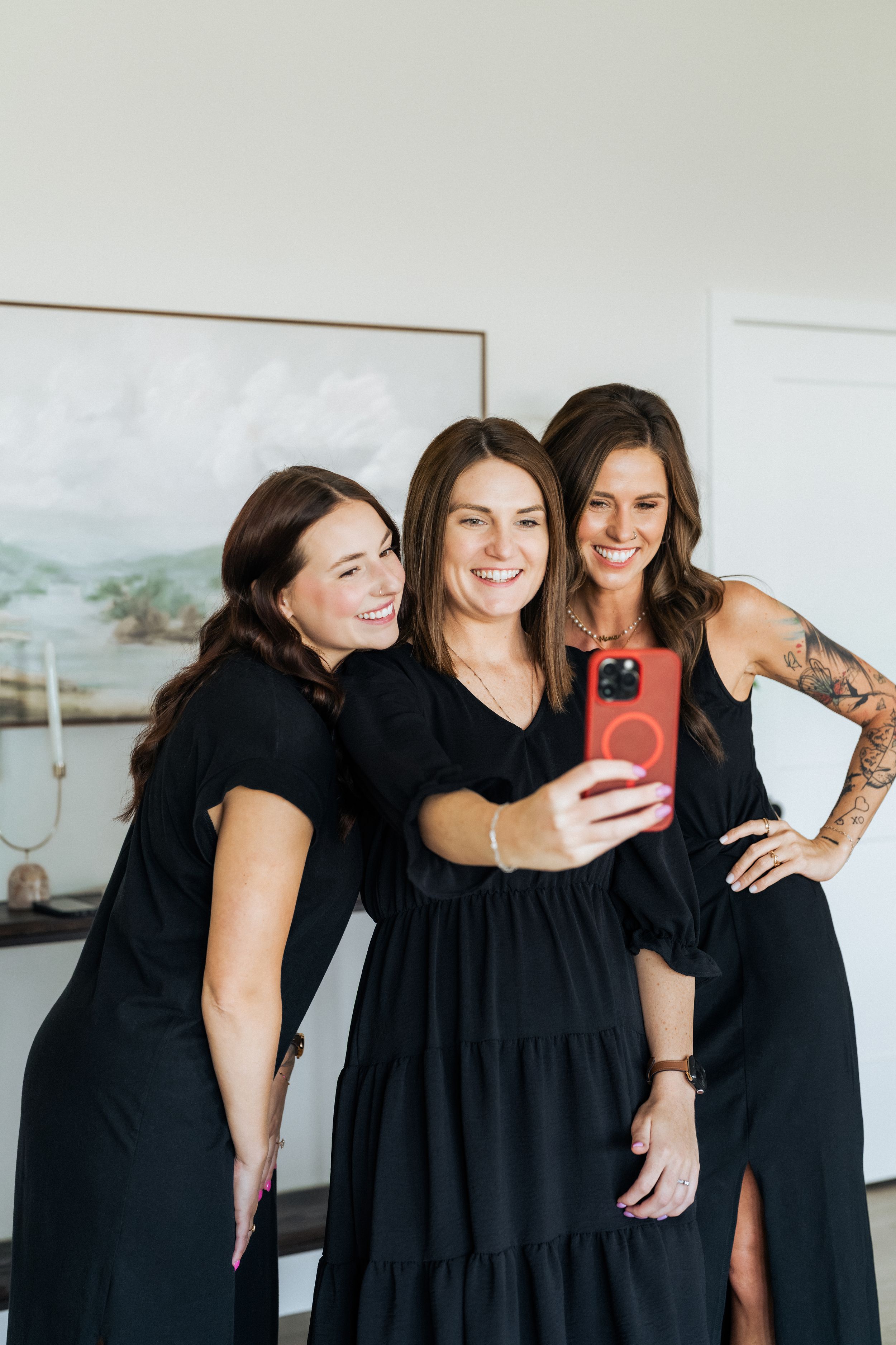 Three women in black dresses smiling and taking a selfie with a red phone indoors.