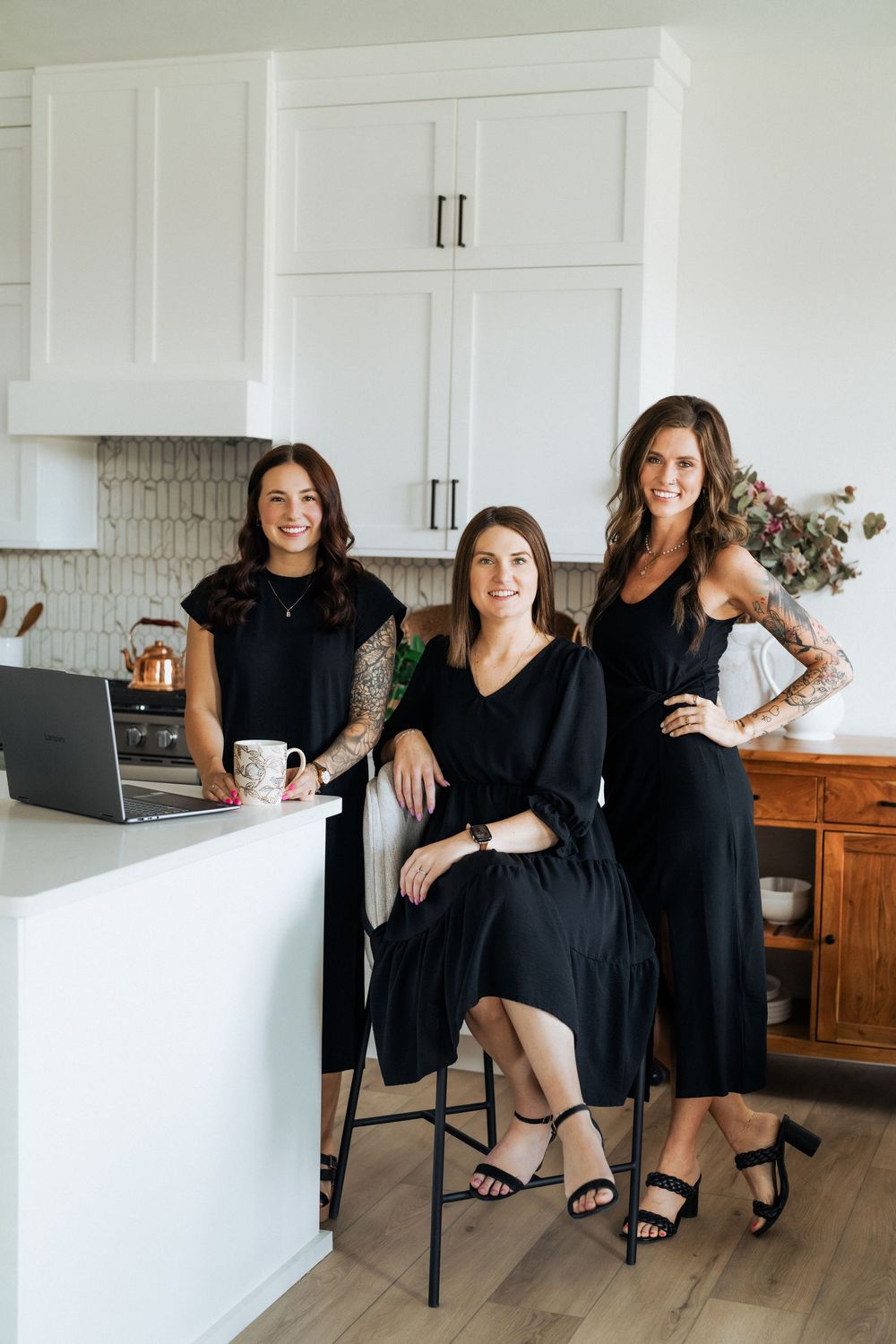 Three women in black dresses smiling in a modern white kitchen with laptop and coffee mug.