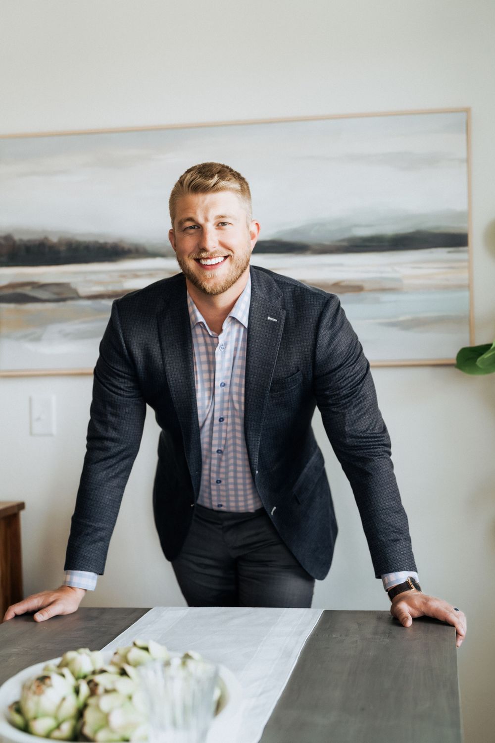 Smiling man in dark blazer leaning on dining table with artichokes, abstract landscape art on wall behind him.