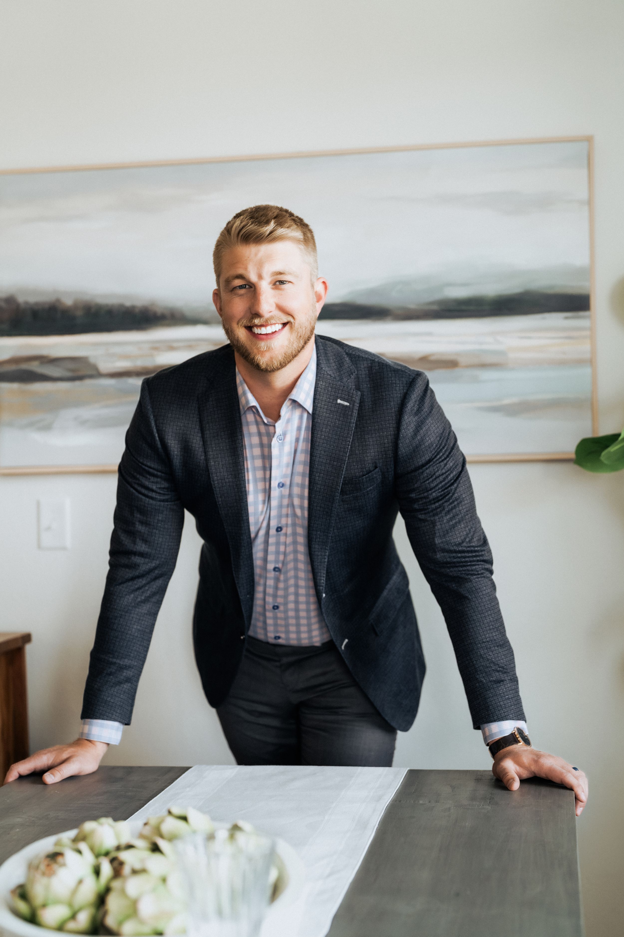 Smiling man in dark blazer leaning on dining table with artichokes, abstract landscape art on wall behind him.