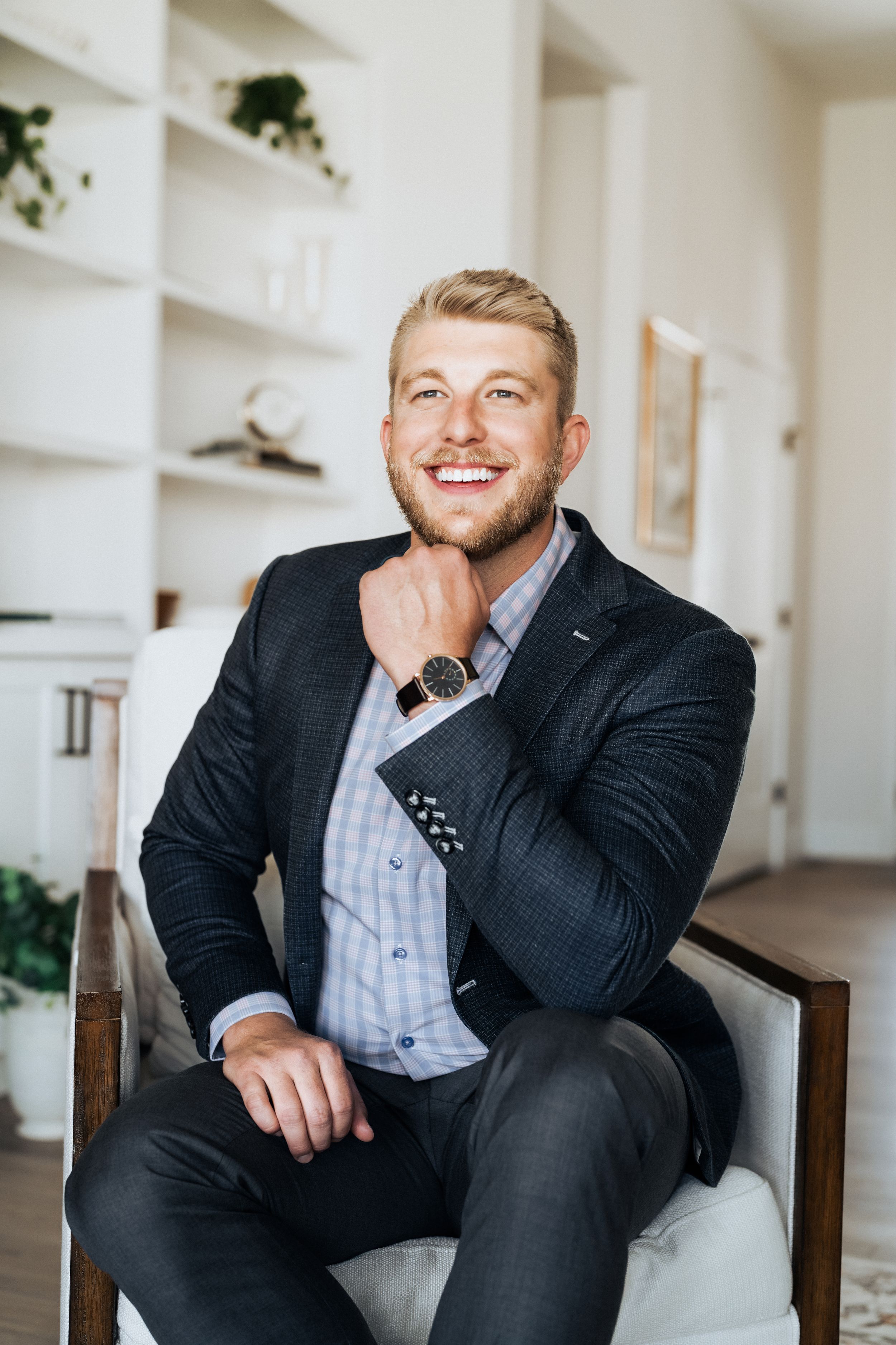 Smiling young businessman in dark blazer and watch sitting confidently in a modern white chair indoors.