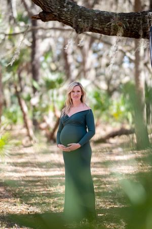 An expecting mother in a teal gown poses gracefully in a sunlit wooded area during a maternity photoshoot.