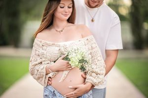 Expecting parents pose outdoors as they hold small white wildflowers against the mother's baby bump in a lace top.