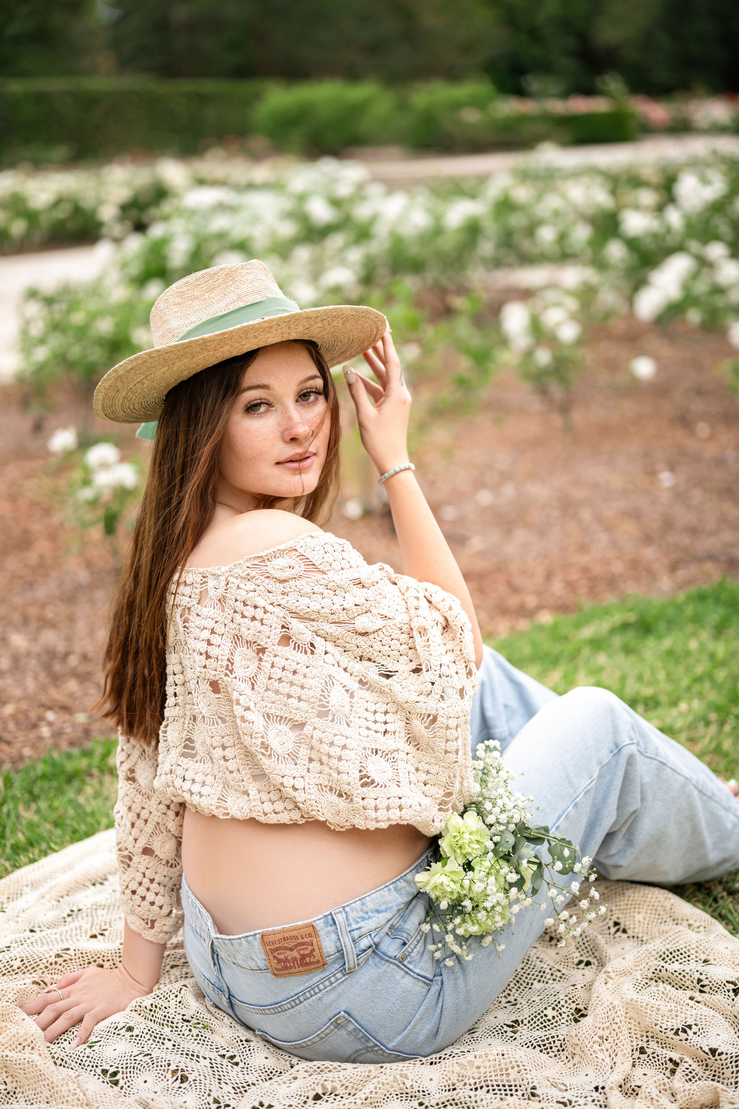 A photoshoot in a garden features a bohemian style crochet top paired with denim and a sun hat amid white flowers.