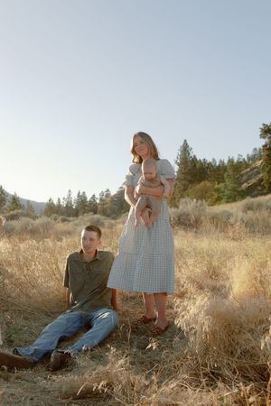 A romantic couple sits together in golden dry grass during a sunny outdoor photo session in gingham dress and casual wear.