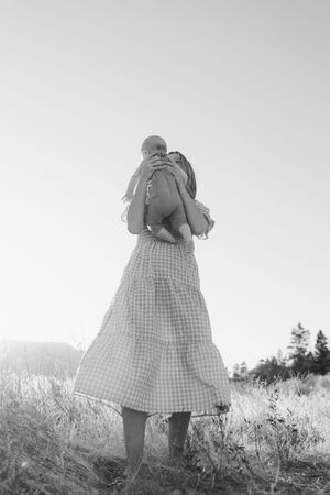 A figure in a gingham dress stands in a meadow looking away from the camera, shown in black and white.