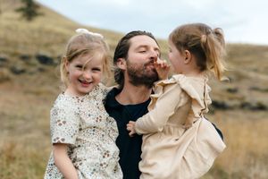 A parent and two young children share joyful smiles and laughter outdoors on a hillside in nature.