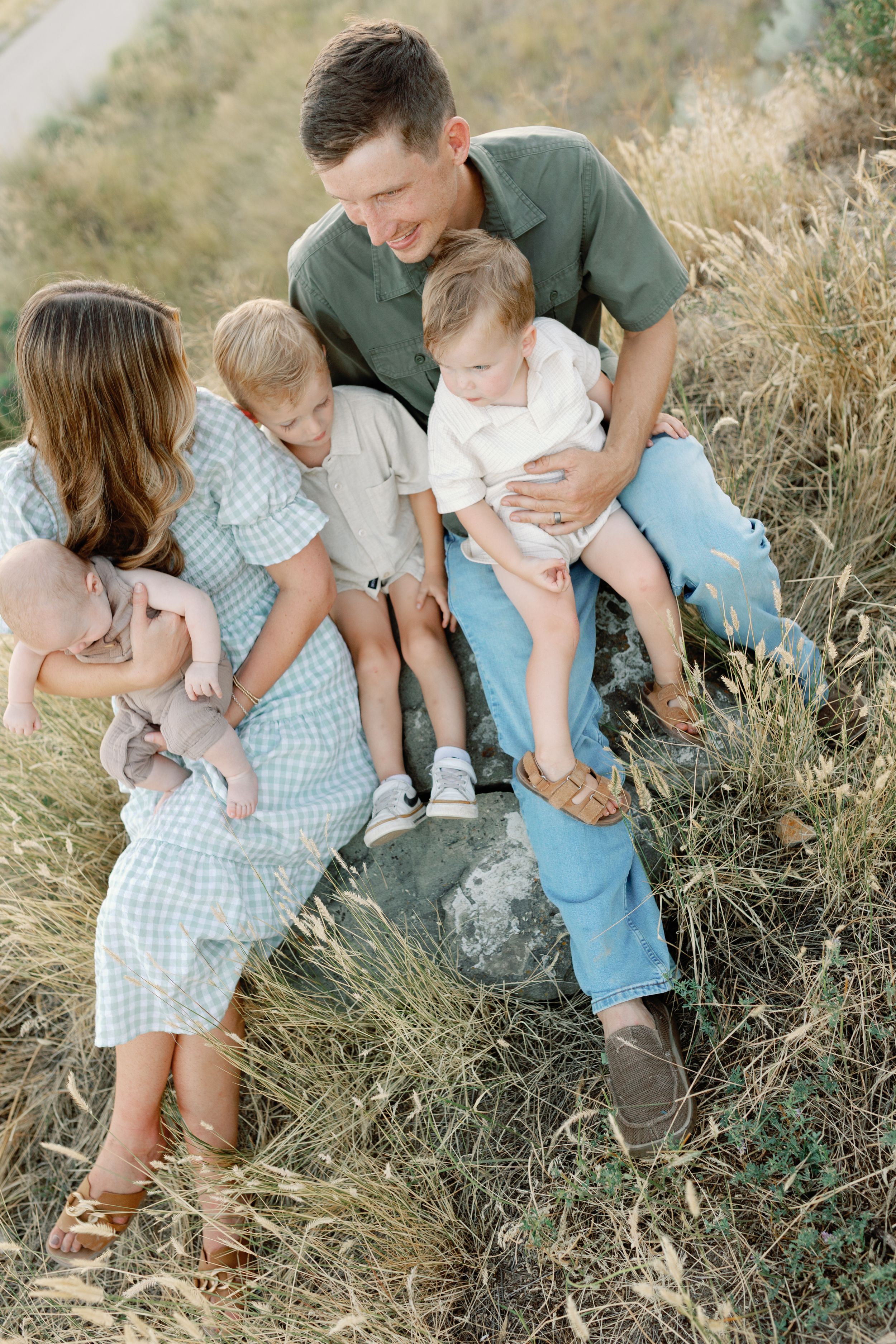 Family sits together on grassy hillside wearing coordinated light blue and green outfits in outdoor portrait session.