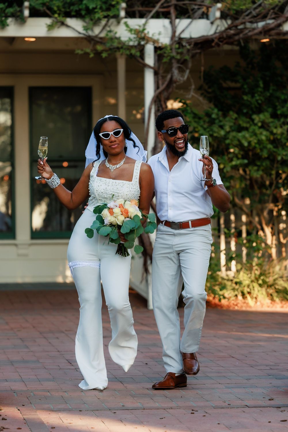 Wedding party members in coordinated white outfits dance and celebrate on a brick patio during sunset.
