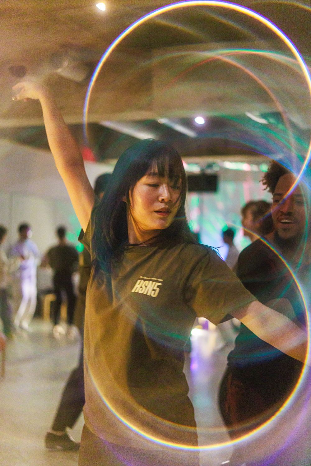 A dancer performs fluid movements under colorful lights creating motion blur trails in a dimly lit indoor space.