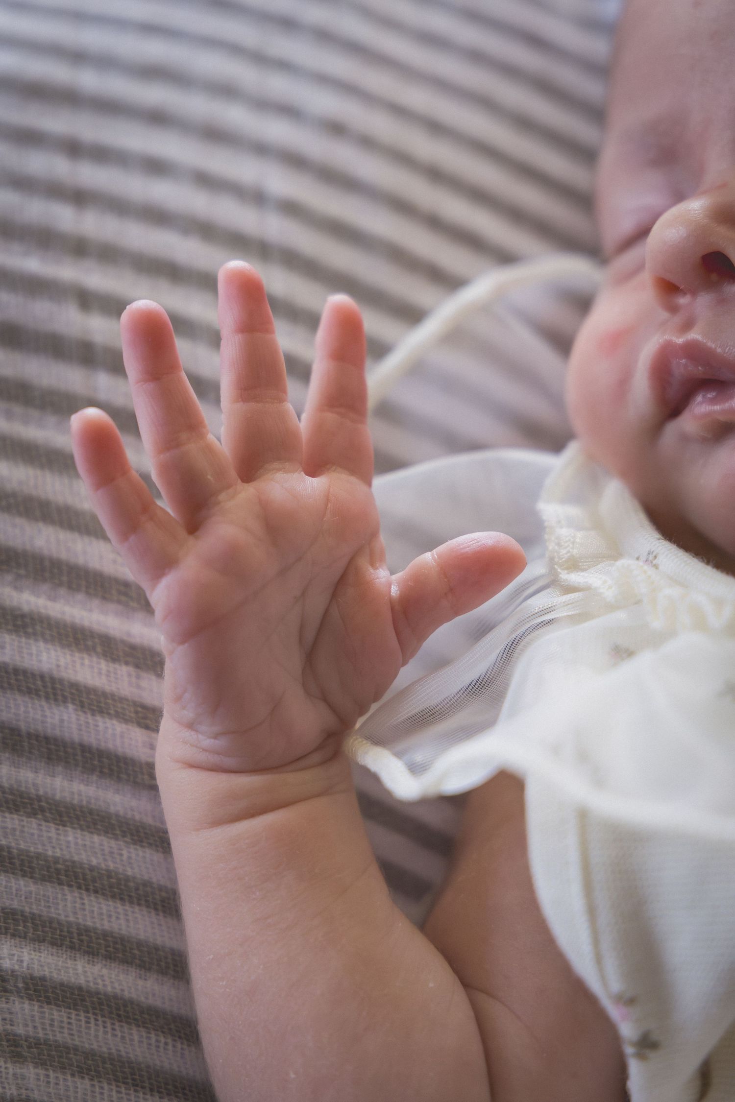 Close-up of a newborn baby's tiny open hand raised up, wearing a white dress, lying on striped fabric.