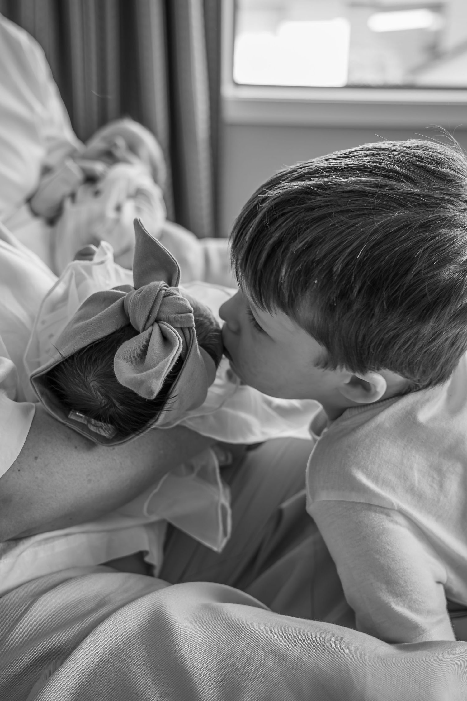 Black and white photo of a young boy gently kissing a newborn baby with a bow, in a tender family moment.