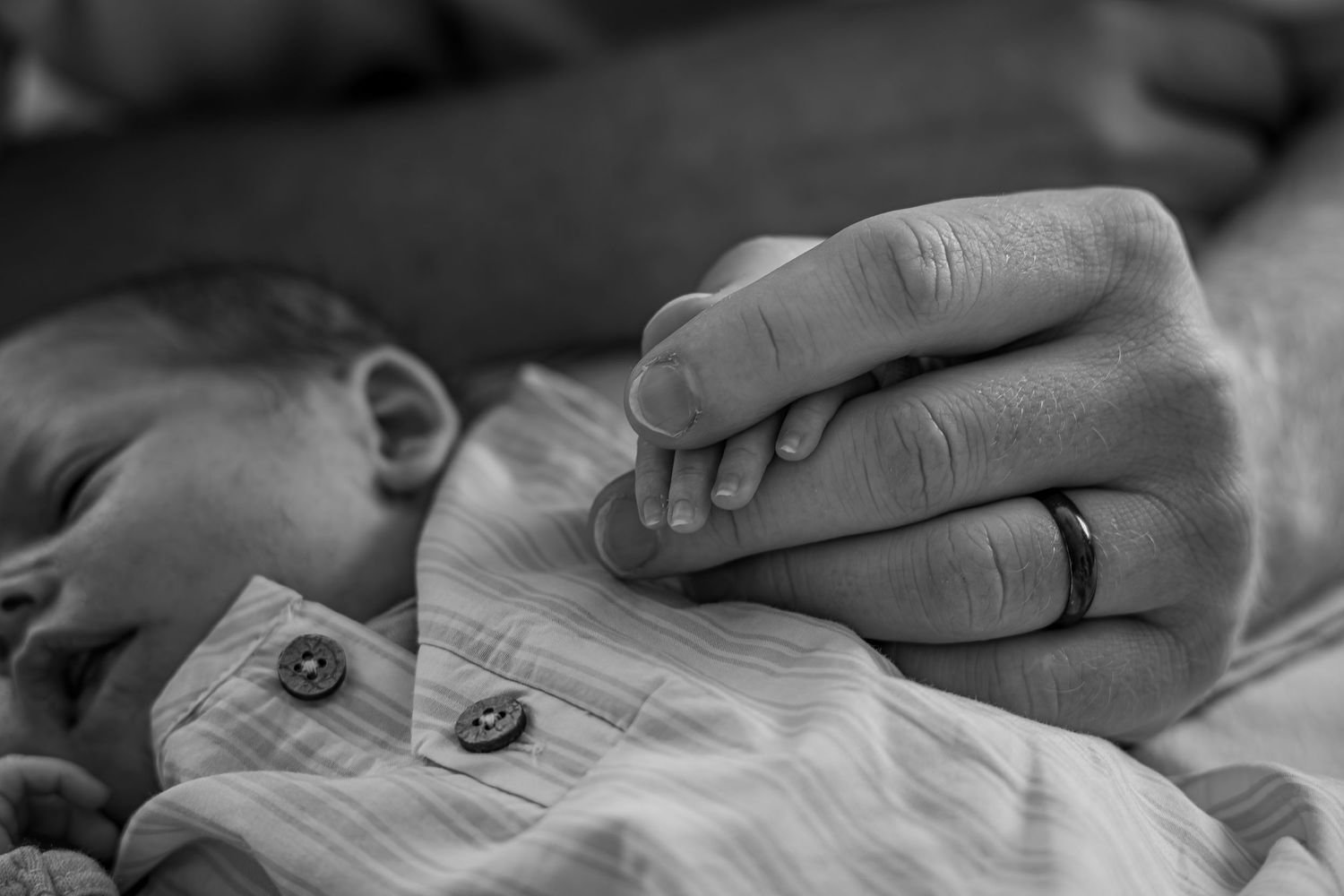 Black and white photo of a newborn baby holding a parent's finger, with a wedding ring visible.