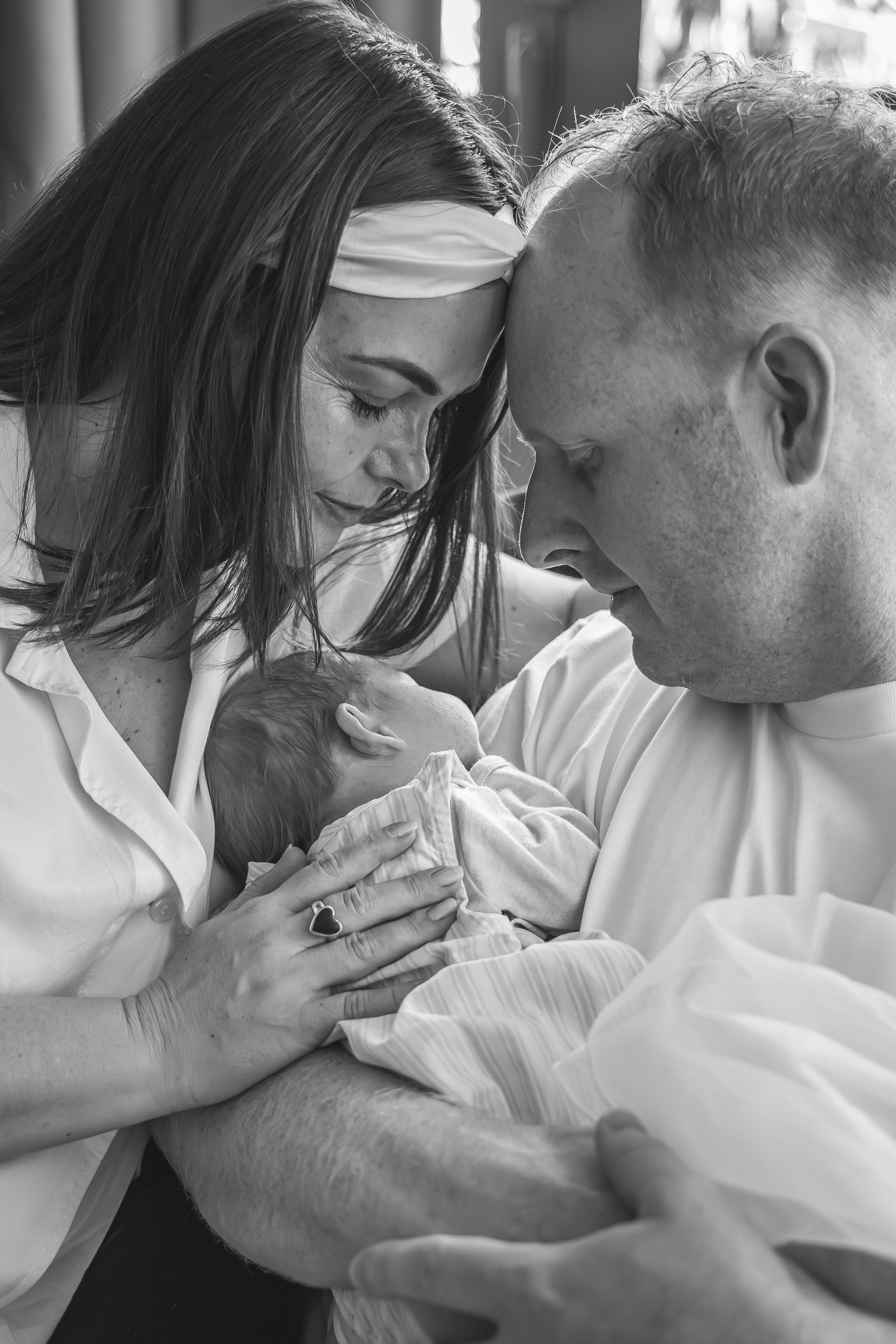 Black and white photo of new parents lovingly gazing at their newborn baby wrapped in white cloth.