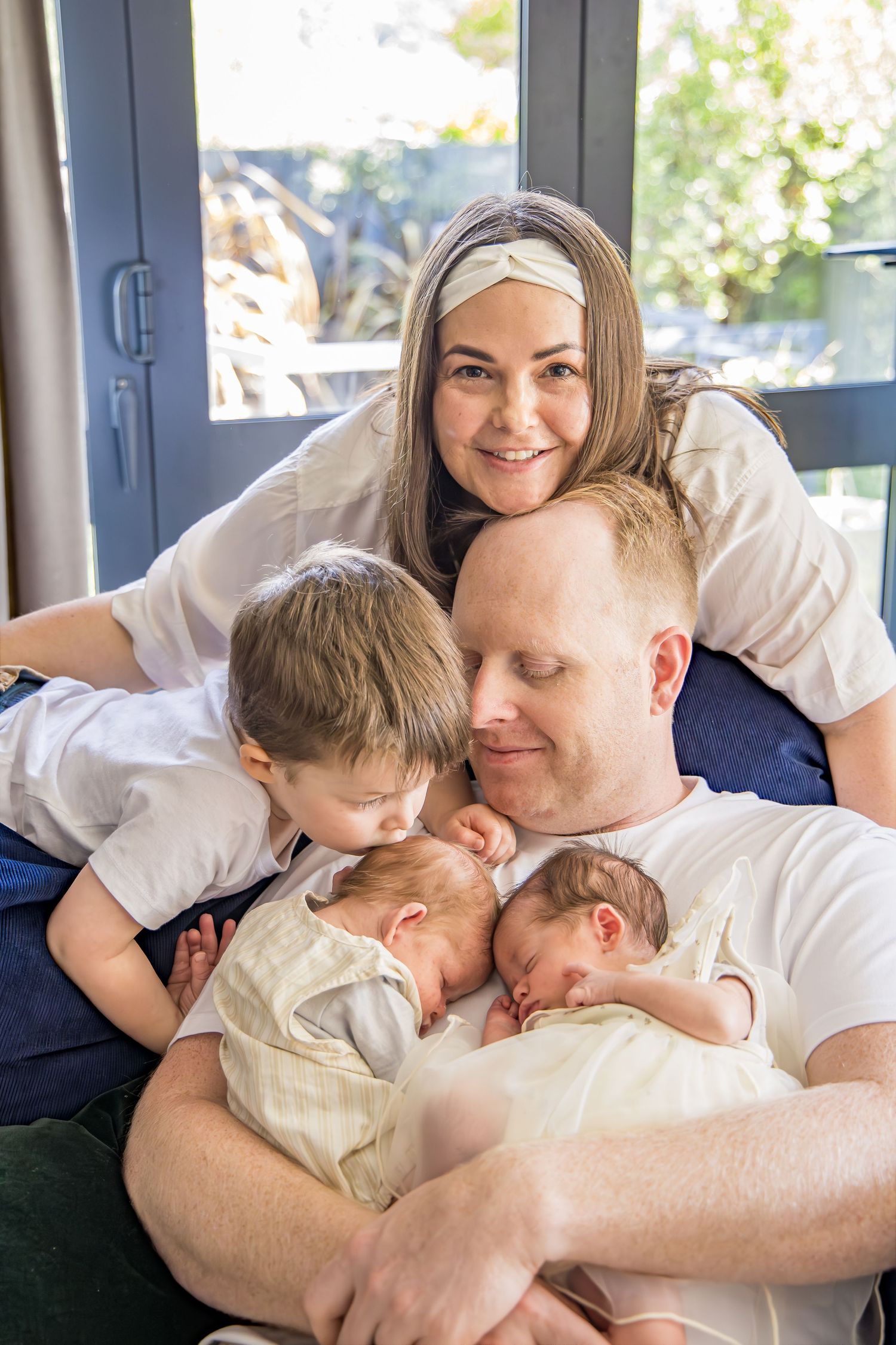 Happy family portrait with parents, toddler, and newborn twins cuddling together indoors.
