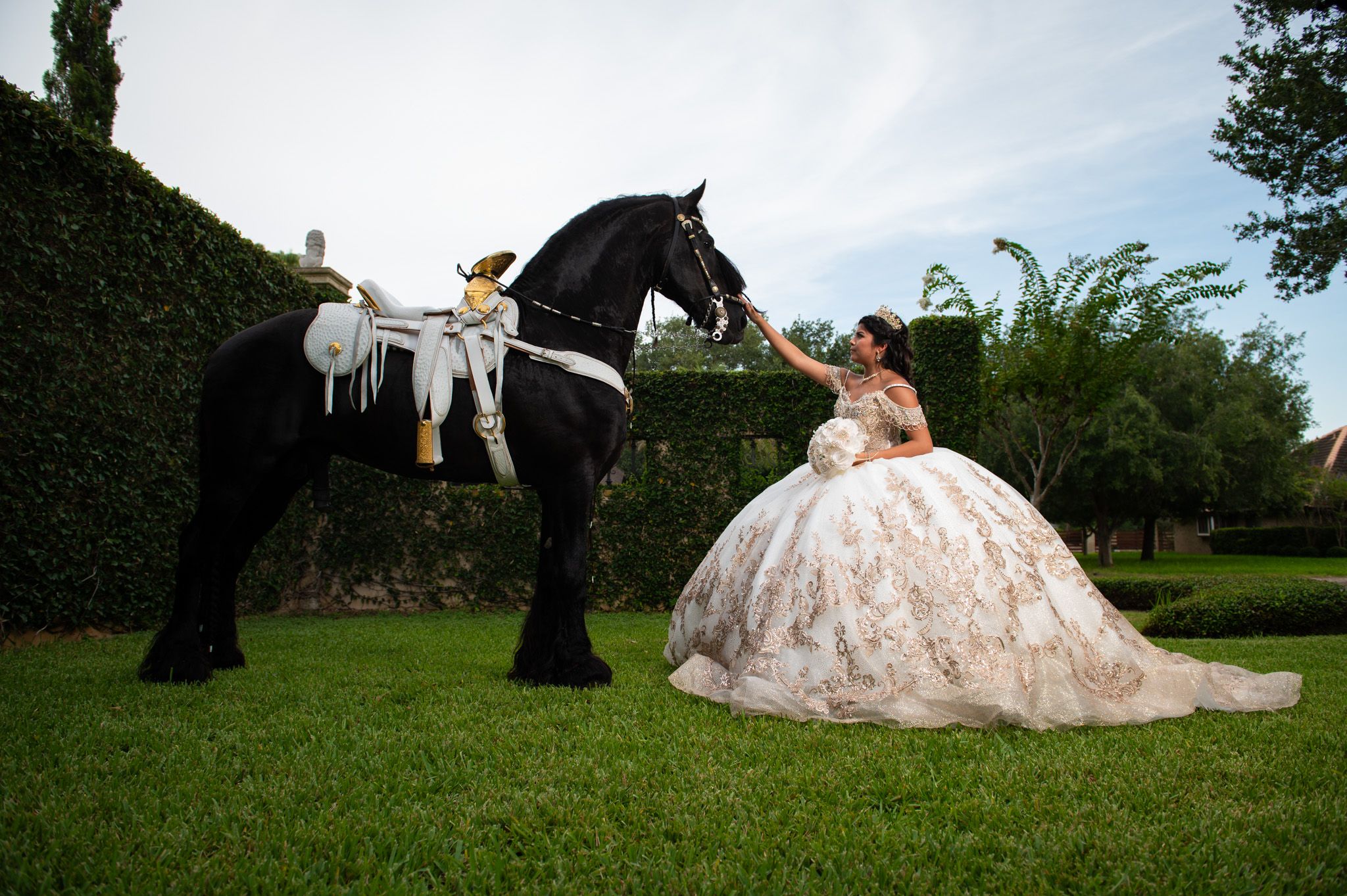 Elegant quinceañera photoshoot featuring a dramatic pose with a majestic black horse in a garden setting.