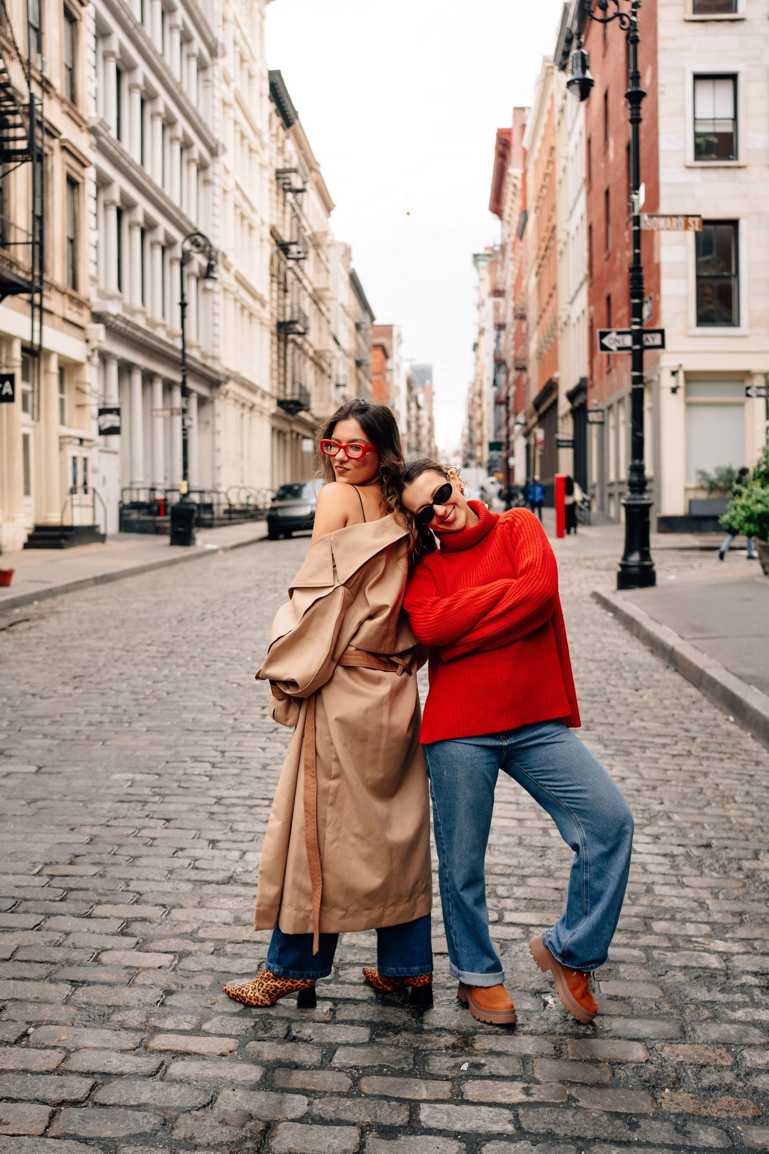 Two people wearing casual outfits dance playfully on a cobblestone street between historic buildings in a city.