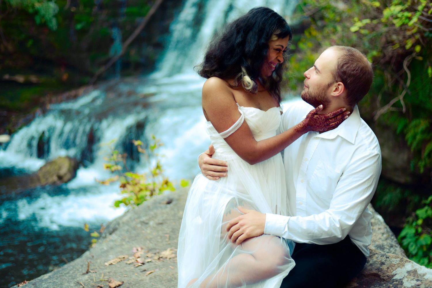 Romantic couple embracing near a waterfall, woman in white dress and man in white shirt with red bow tie.