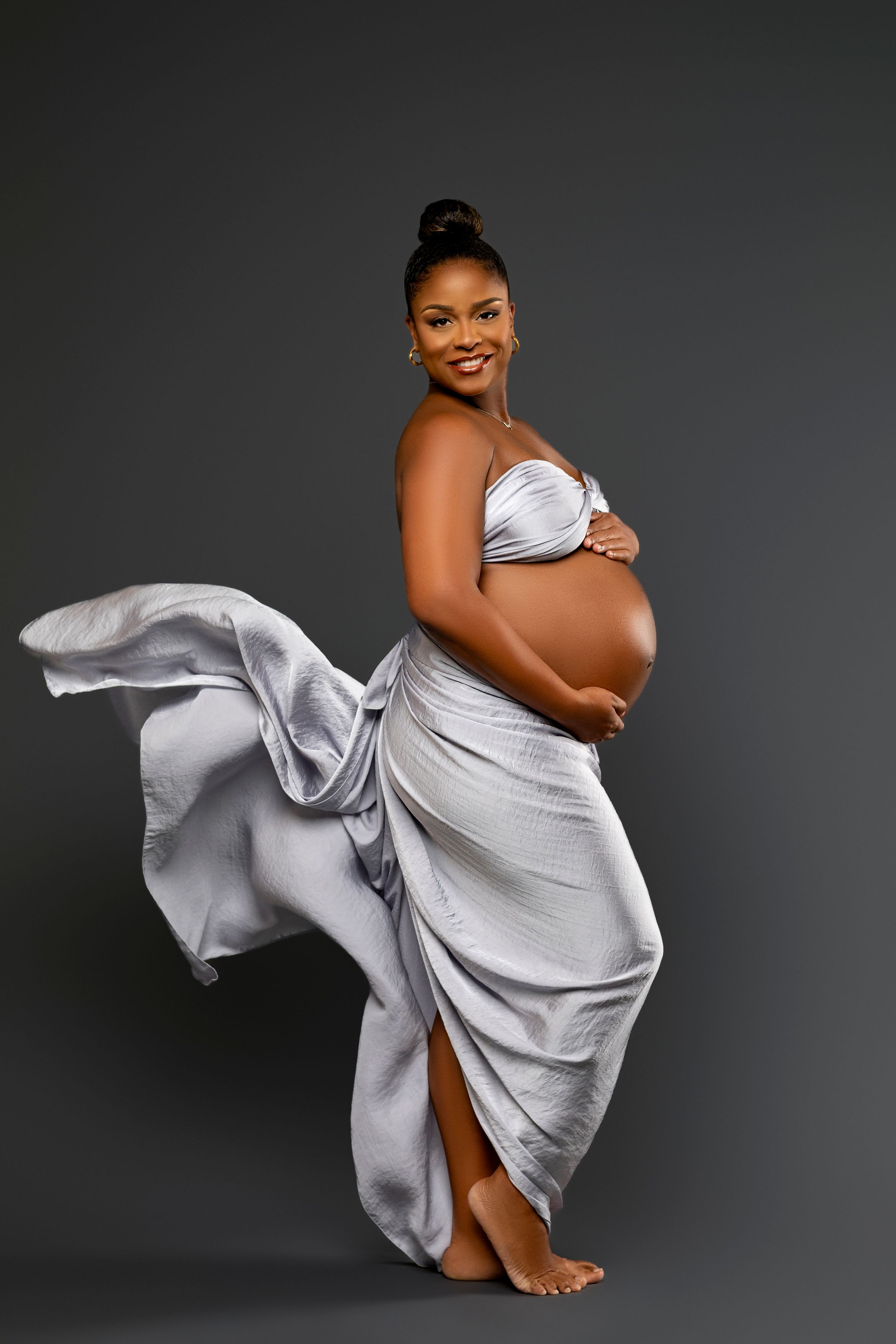 Artistic maternity photo of a woman in flowing silver silk dress against dark gray background showing her pregnant belly.
