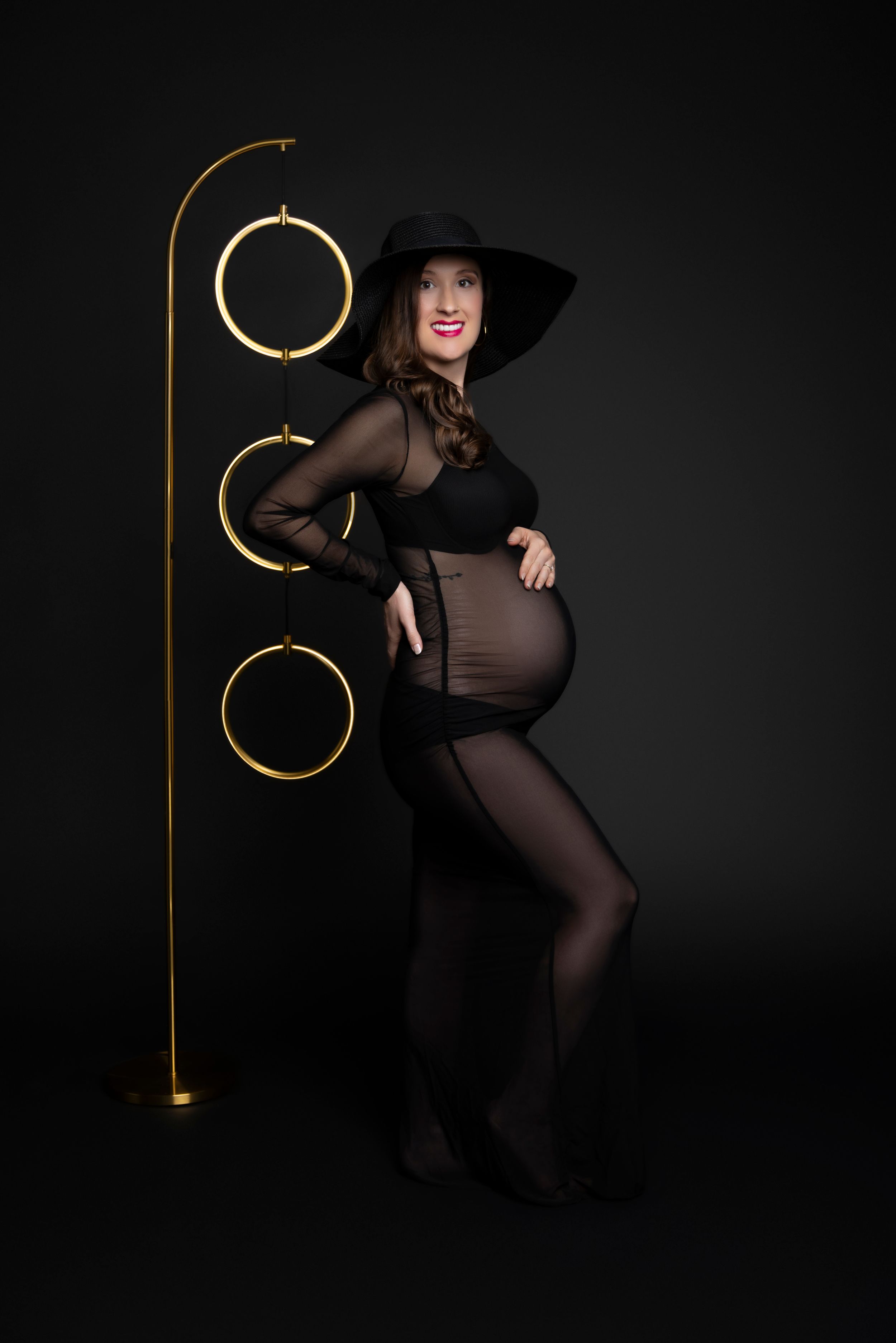 Artistic maternity photo on black background with gold rings display stand and elegant black dress and hat.