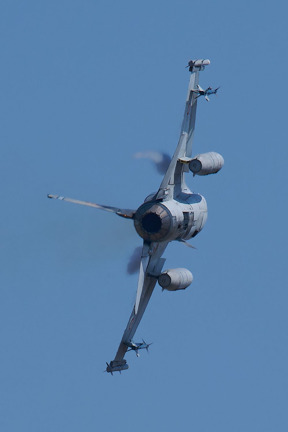Military fighter jet performs a banking maneuver against clear blue sky.