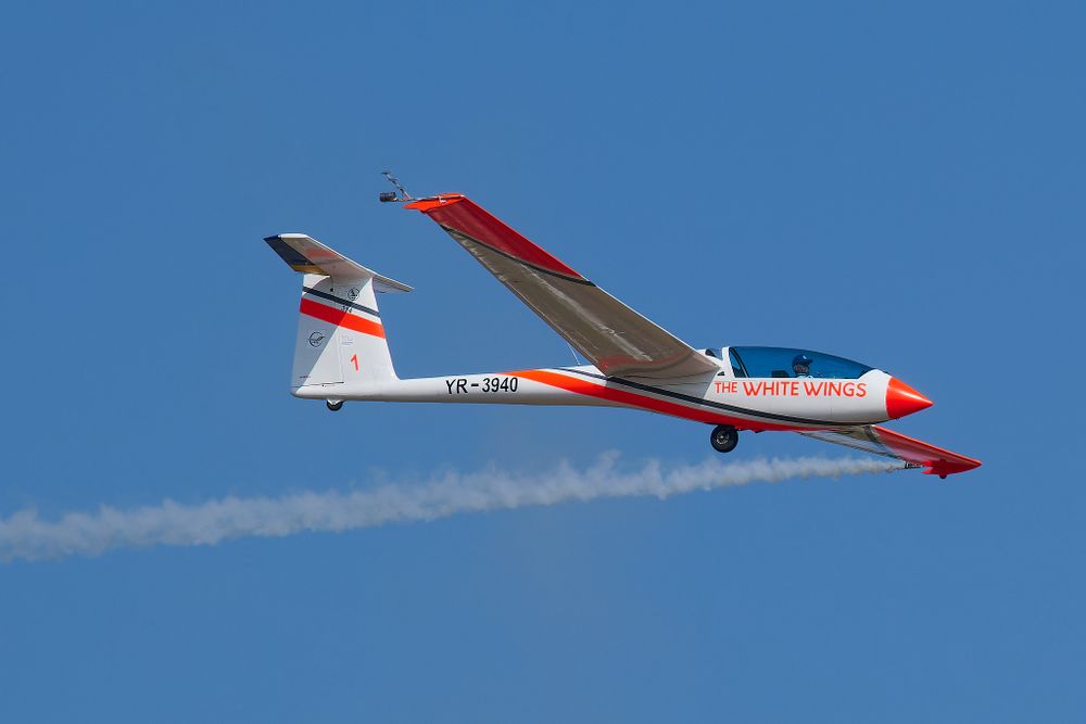 White glider aircraft with red accents releasing smoke trail during flight demonstration.