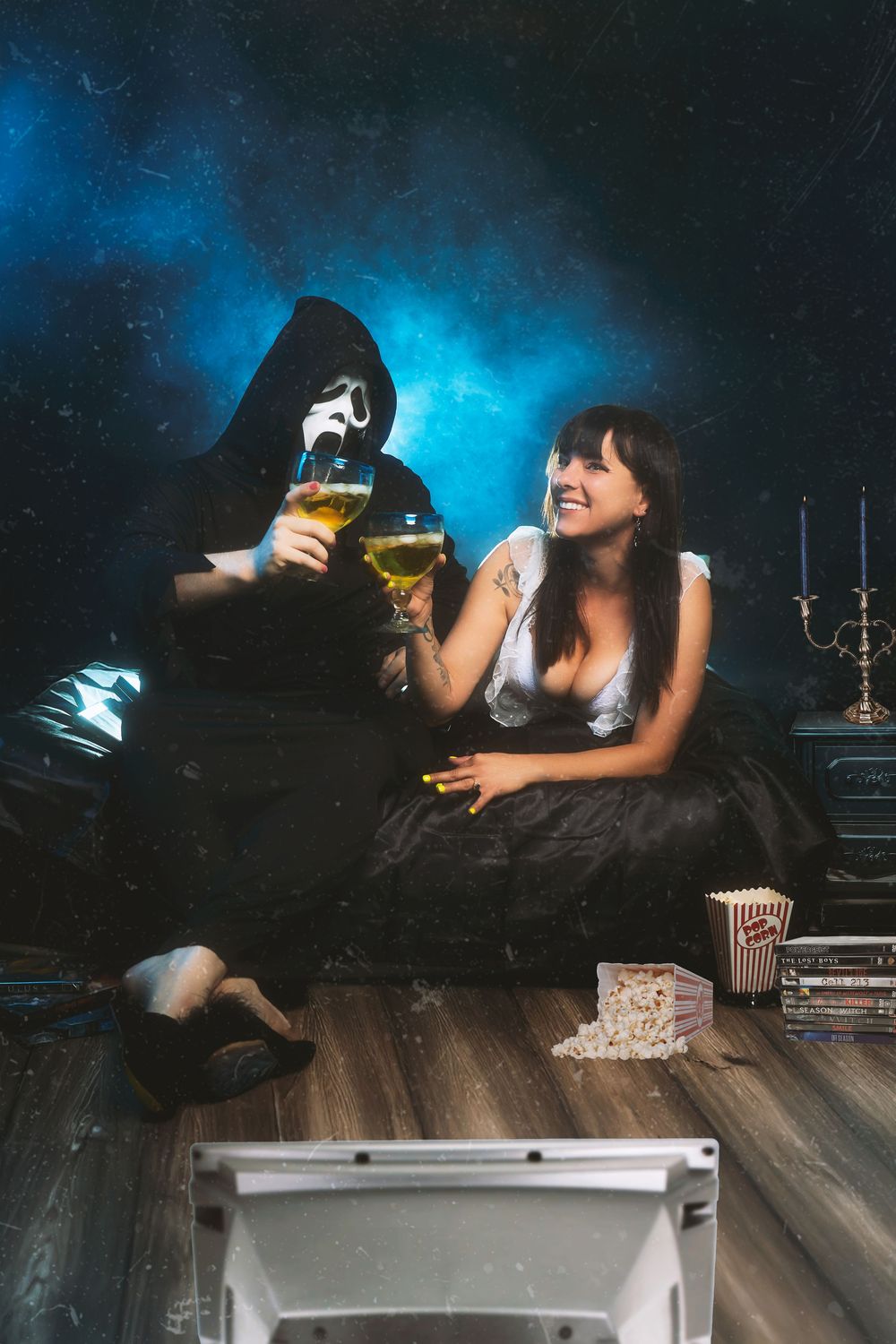 Couple watching TV on floor with popcorn, woman smiling, man in Scream costume, clinking beer glasses in dark room.