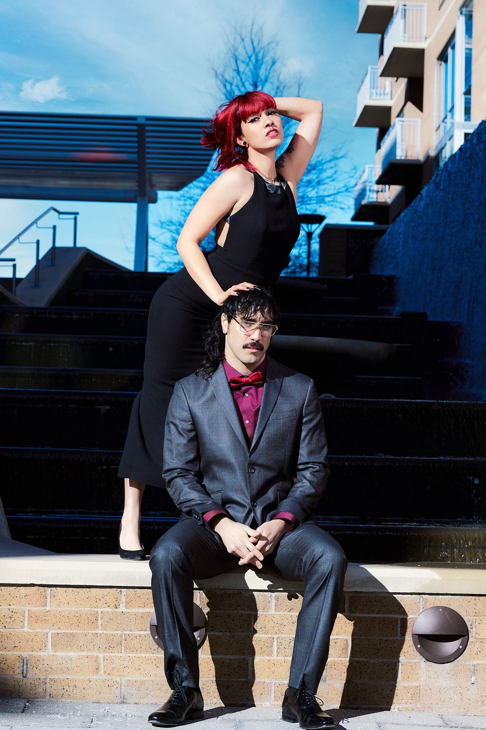 A woman with red hair in a black dress poses above a suited man on outdoor steps in an urban setting.