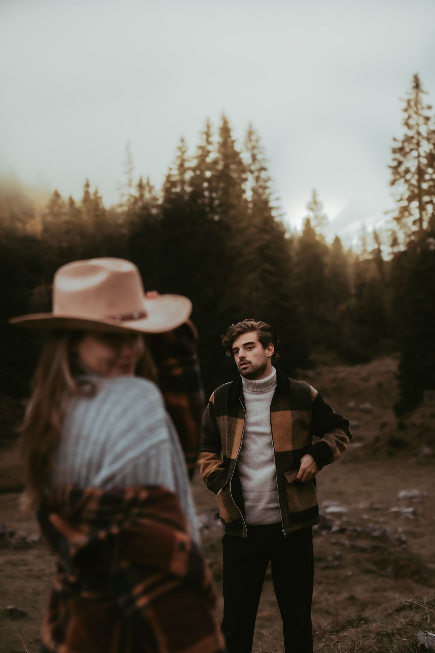 Two people in casual outdoor wear stand on a mountain trail with evergreen forest backdrop.