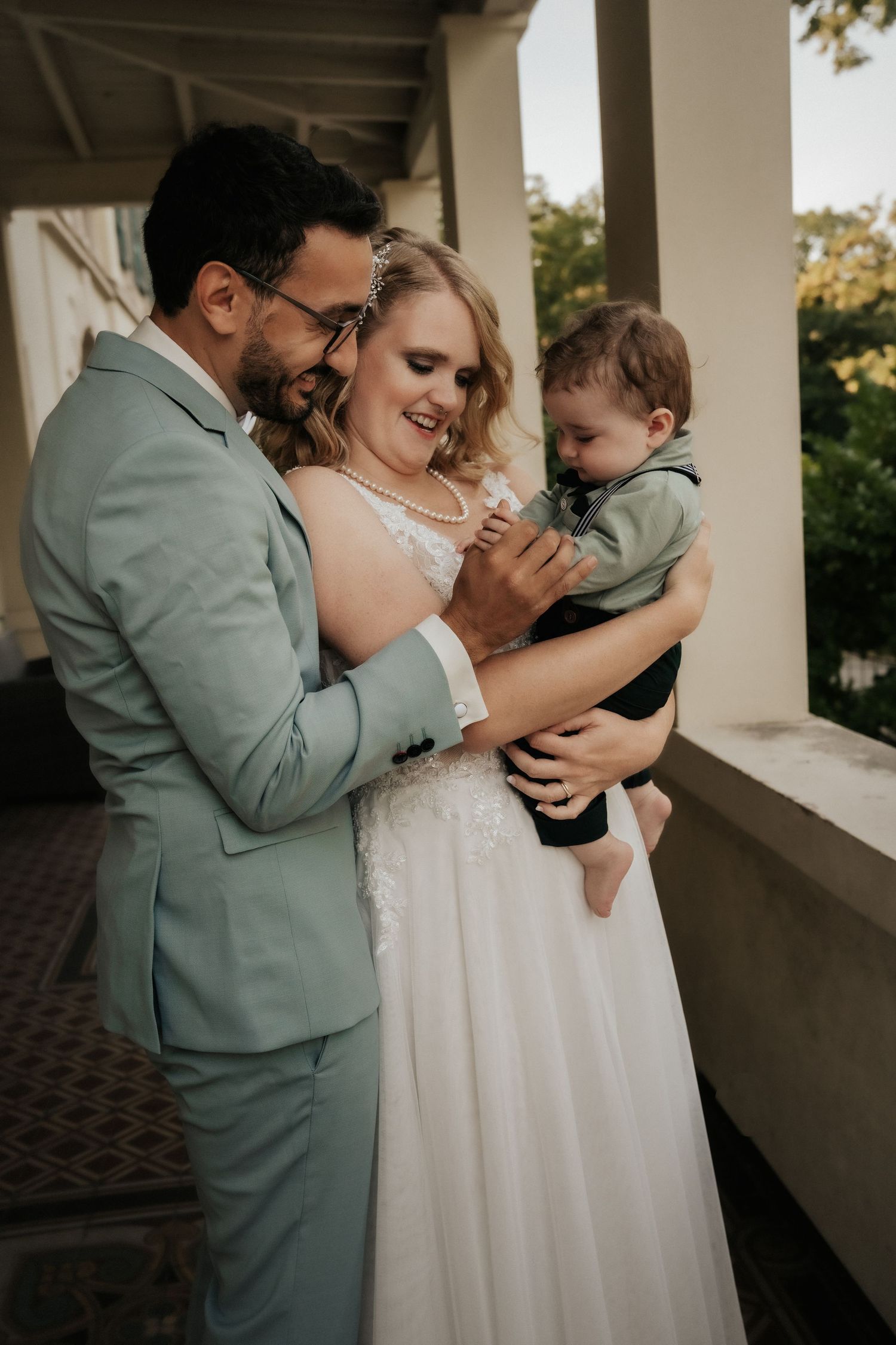 Wedding couple in formal attire share moment with young child on covered porch.