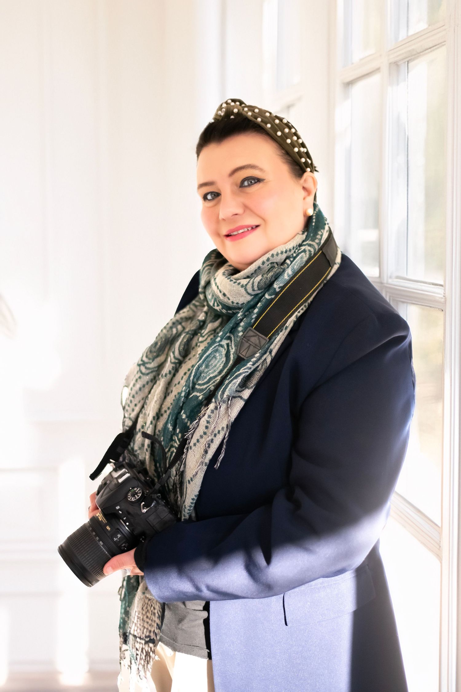 Professional photographer wearing a navy coat and patterned scarf stands by a bright window holding a DSLR camera.