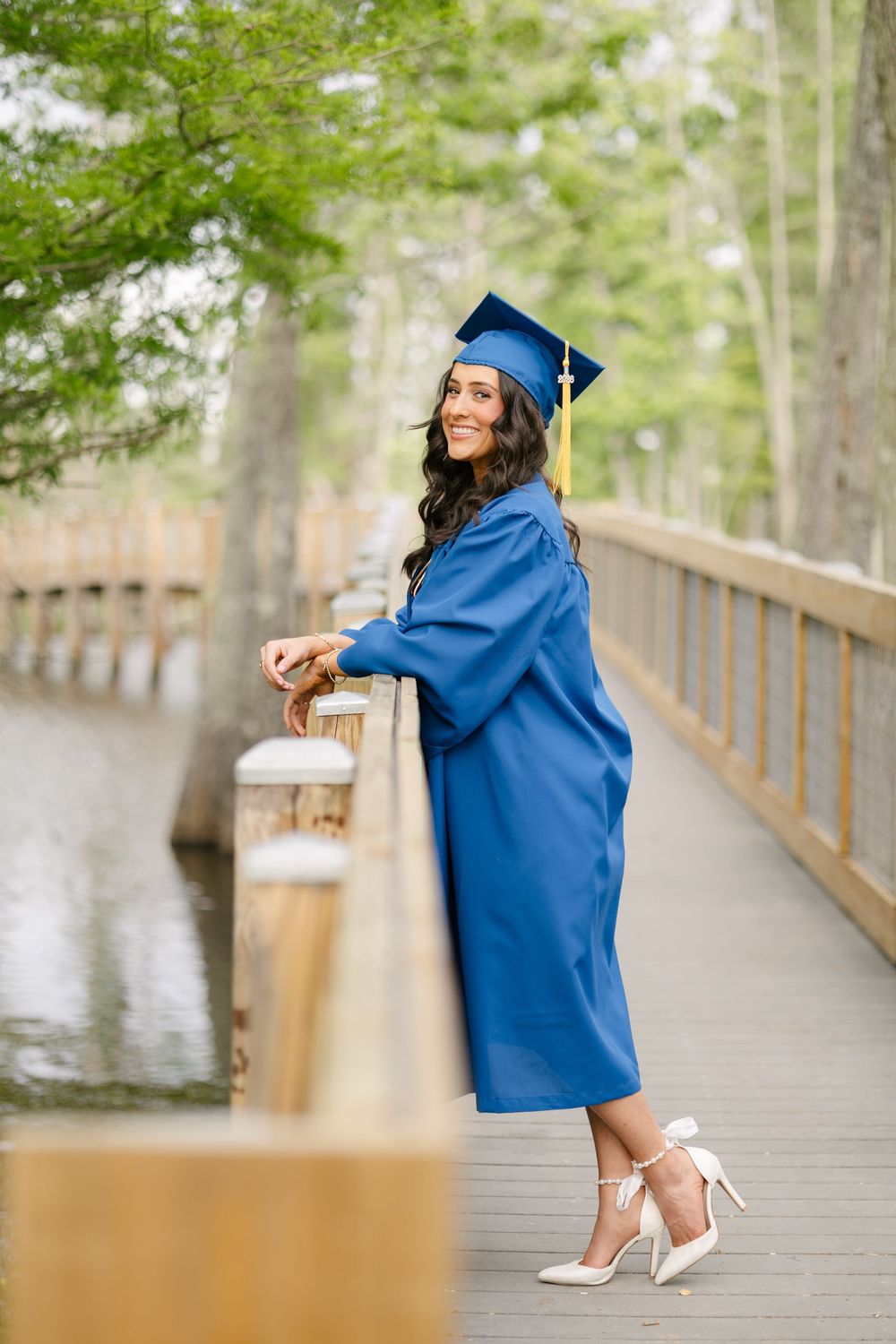 Smiling graduate in blue cap and gown posing on wooden bridge near water, wearing white heels.